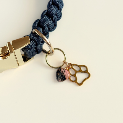 Crystal for dogs pet collar charm featuring a 10mm rhodonite heart shaped bead and a brass gold coloured paw print tag. Pictured attached to a black paracord dog collar.