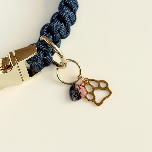 Crystal for dogs pet collar charm featuring a 10mm rhodonite heart shaped bead and a brass gold coloured paw print tag. Pictured attached to a black paracord dog collar.