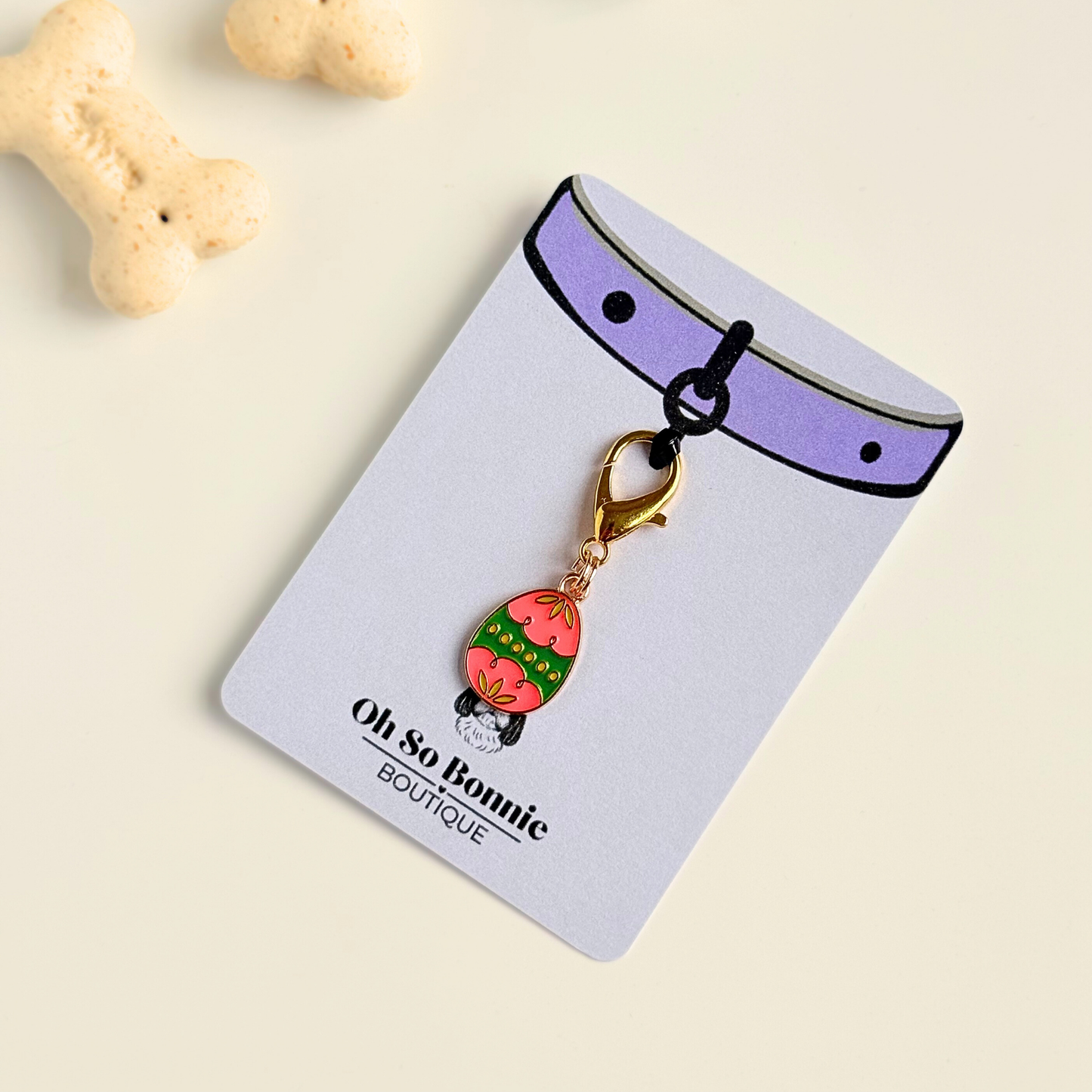 Colourful pink and green patterned Easter egg enamel dog collar charm that easily attaches to collars and harnesses with a gold plated lobster clasp. Pictured on the display card next to some dog biscuits.￼