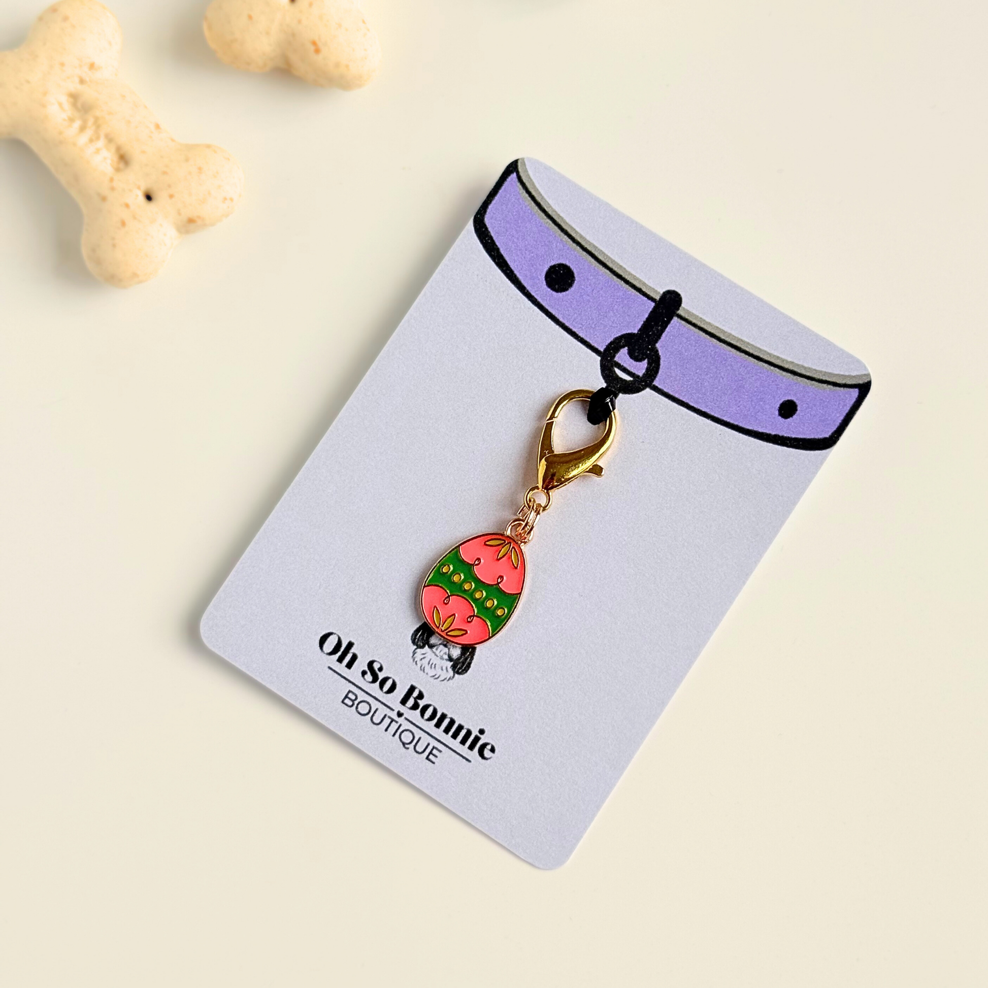 Colourful pink and green patterned Easter egg enamel dog collar charm that easily attaches to collars and harnesses with a gold plated lobster clasp. Pictured on the display card next to some dog biscuits.￼