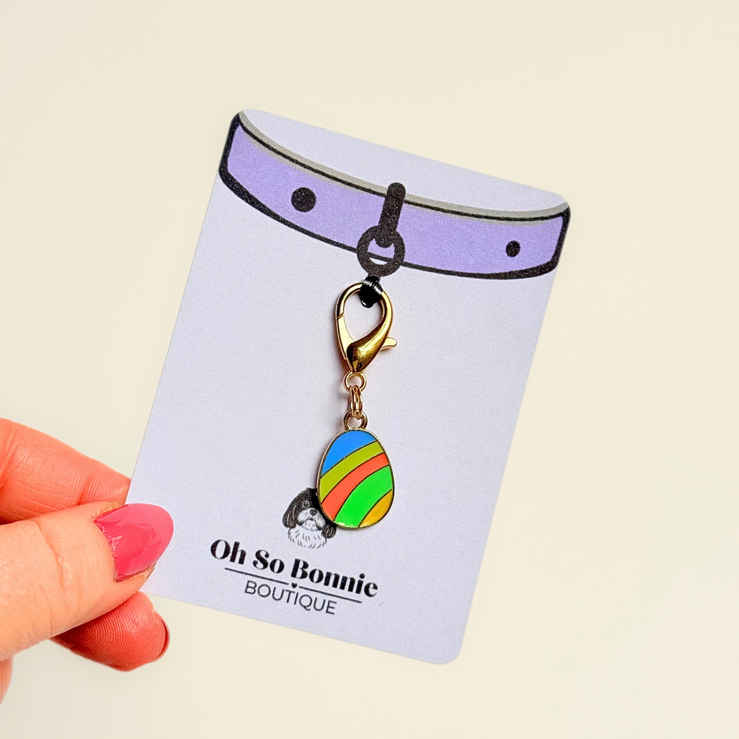 Colourful striped Easter egg enamel dog collar charm that easily attaches to collars and harnesses with a gold plated lobster clasp. Pictured being held on the display card.￼