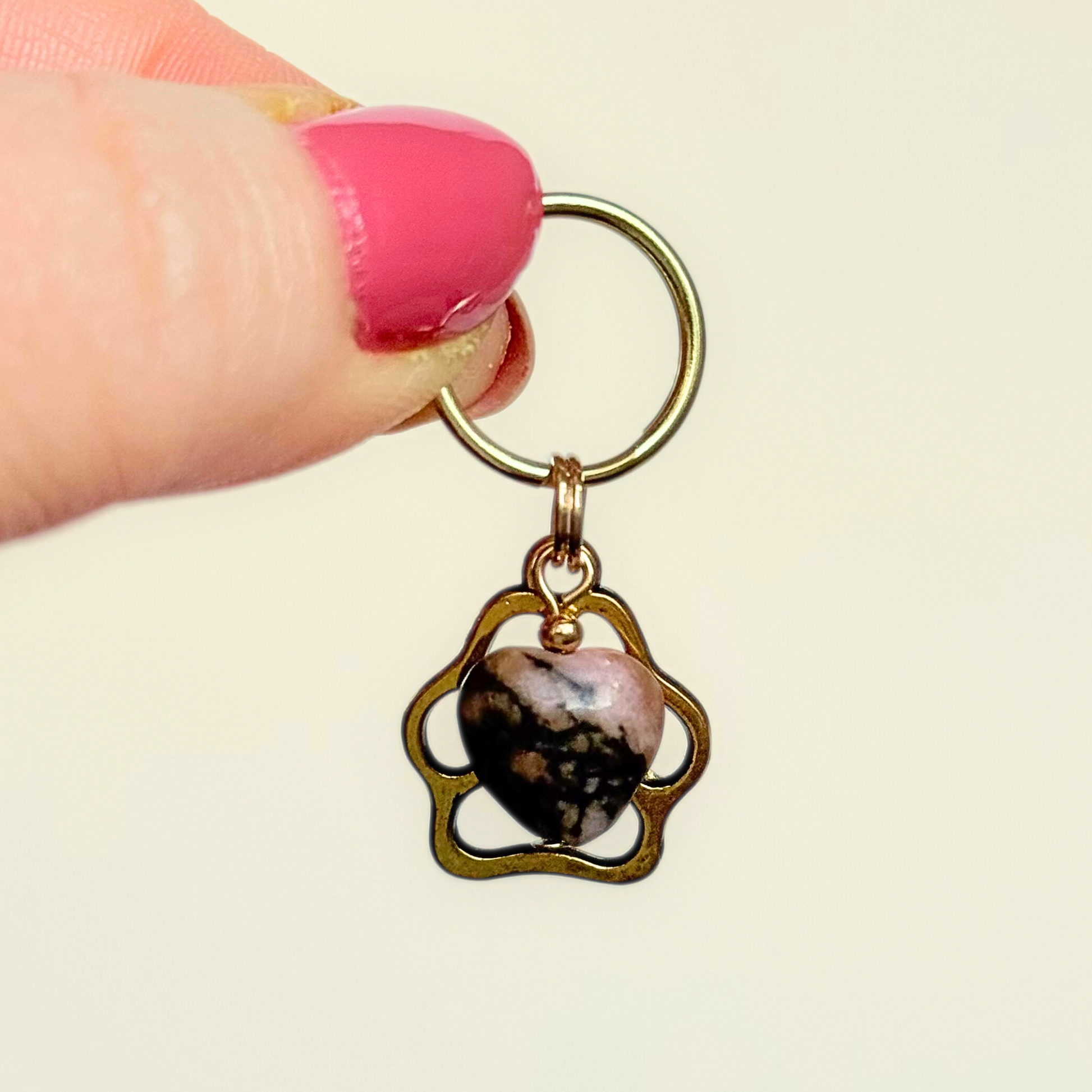 Crystal for dogs pet collar charm featuring a 10mm rhodonite heart shaped bead and a brass gold coloured paw print tag. Pictured being held against a cream background.￼