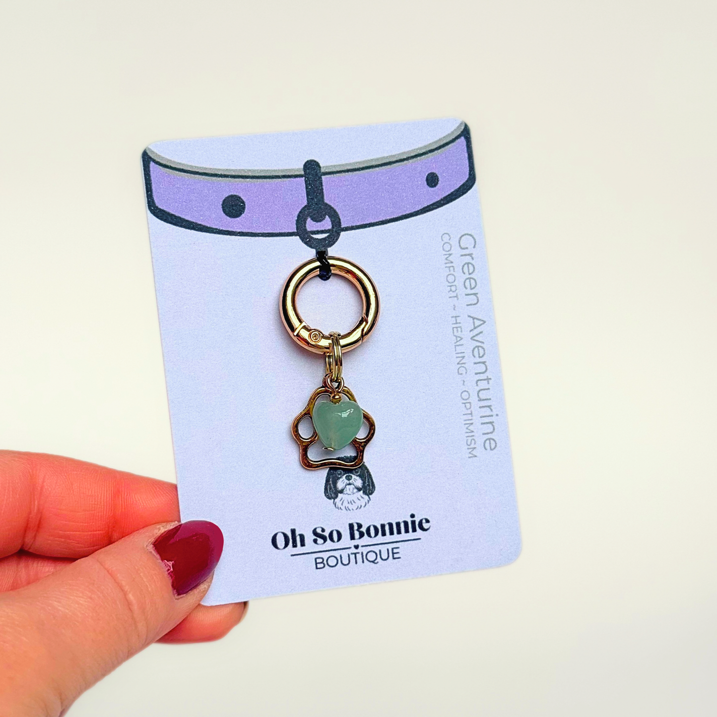 Crystal for dogs pet collar charm featuring a 10mm green aventurine heart shaped bead and a brass gold coloured paw print tag. A stone for comfort, healing and optimism. Pictured on the display card being held up against a cream background.