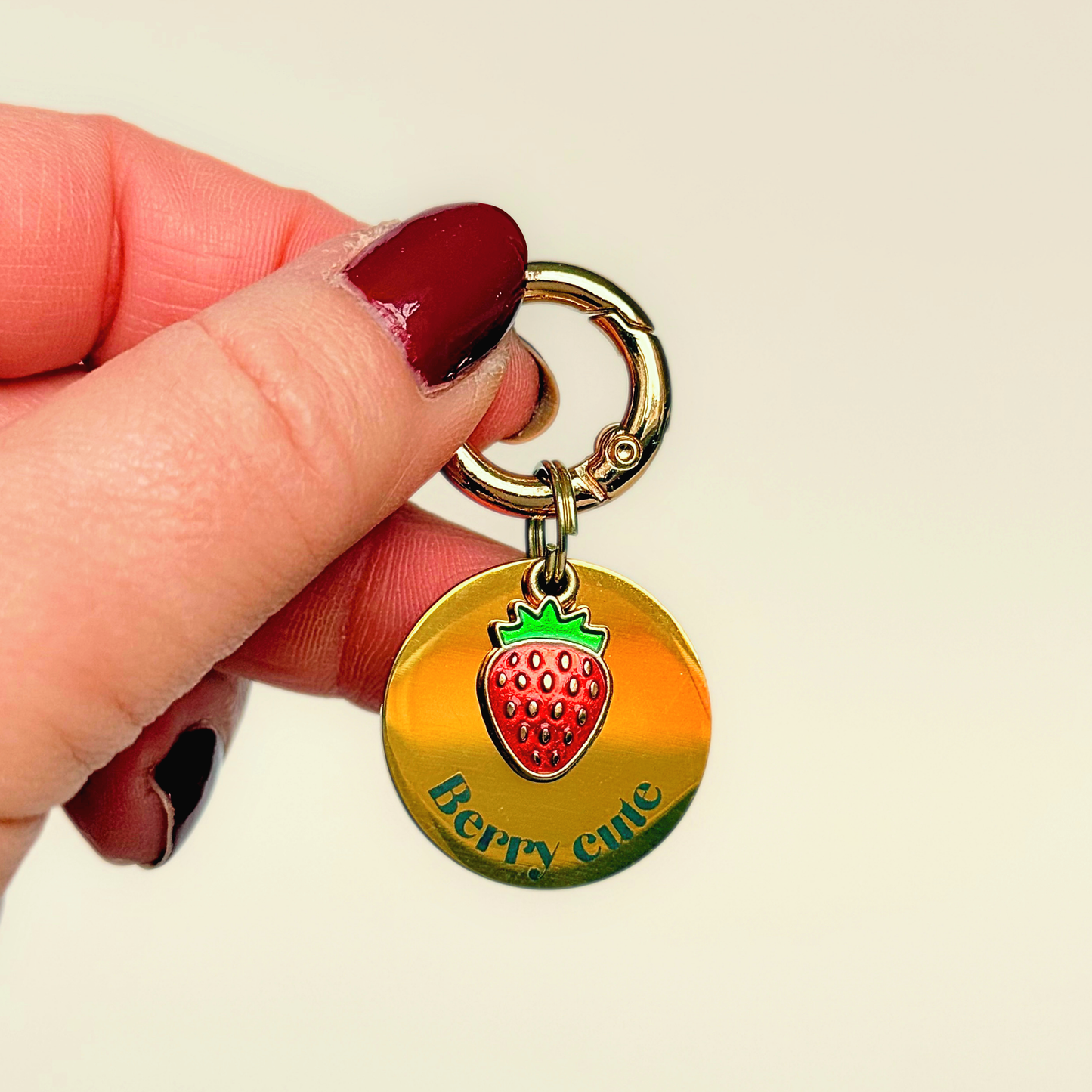 Gold red strawberry dog collar ID tag, Enamel fruit charm with dog name and address details. easily clips onto collar or harness. Shown being held against a cream background.