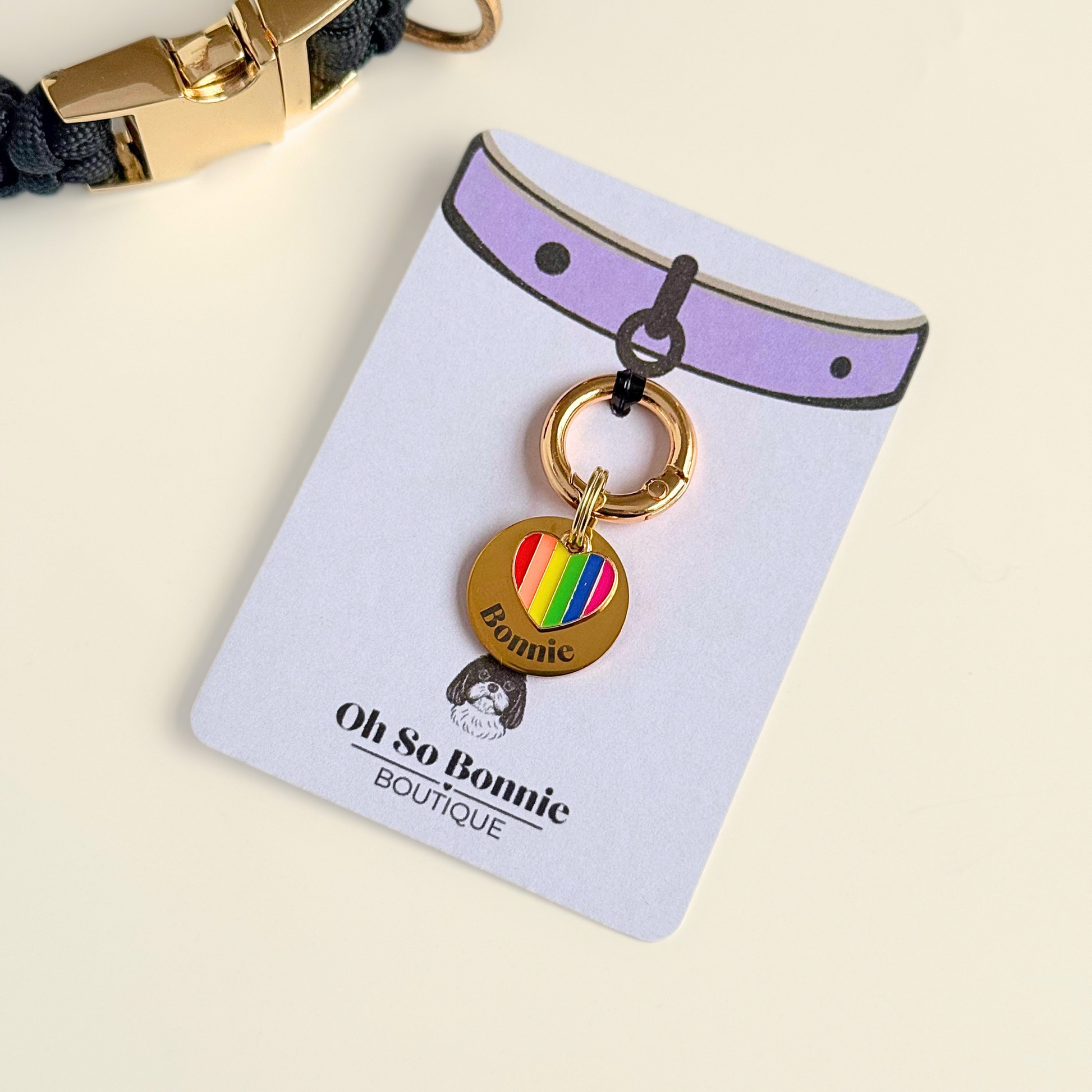 Gold brass rainbow striped heart dog collar ID tag, Enamel charm with dog name and address details. easily clips onto collar or harness. Pictured on the display card next to a black paracord dog collar.