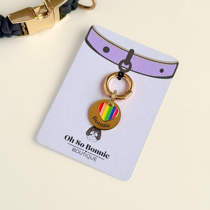 Gold brass rainbow striped heart dog collar ID tag, Enamel charm with dog name and address details. easily clips onto collar or harness. Pictured on the display card next to a black paracord dog collar.