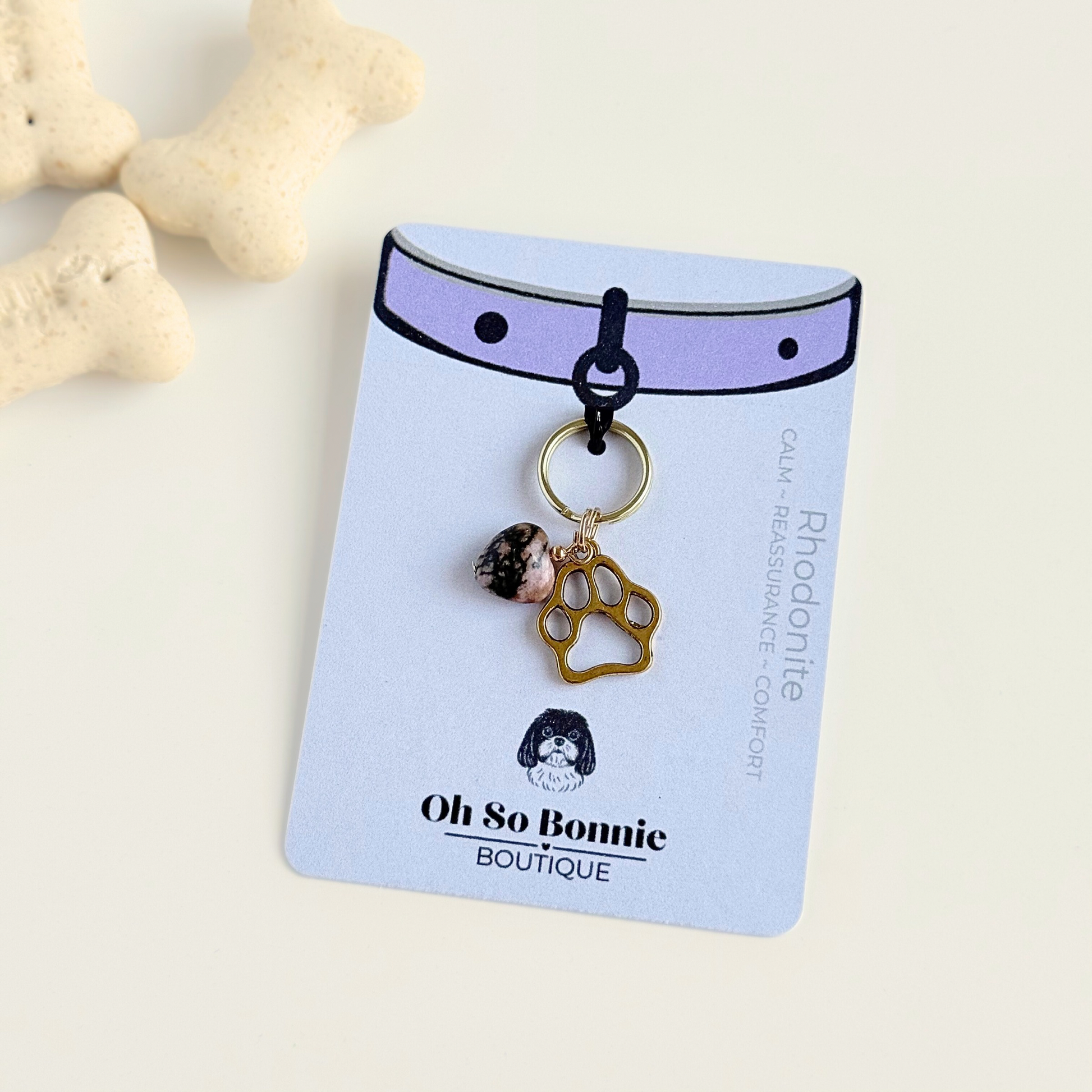 Crystal for dogs pet collar charm featuring a 10mm rhodonite heart shaped bead and a brass gold coloured paw print tag. On display card next to dog biscuits.