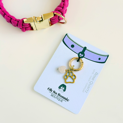 Crystal for dogs pet collar charm featuring a 10mm rose quartz heart shaped bead and a brass gold coloured paw print tag. A stone for love, calm and comfort. Pictured on the display card next to a pink collar.
