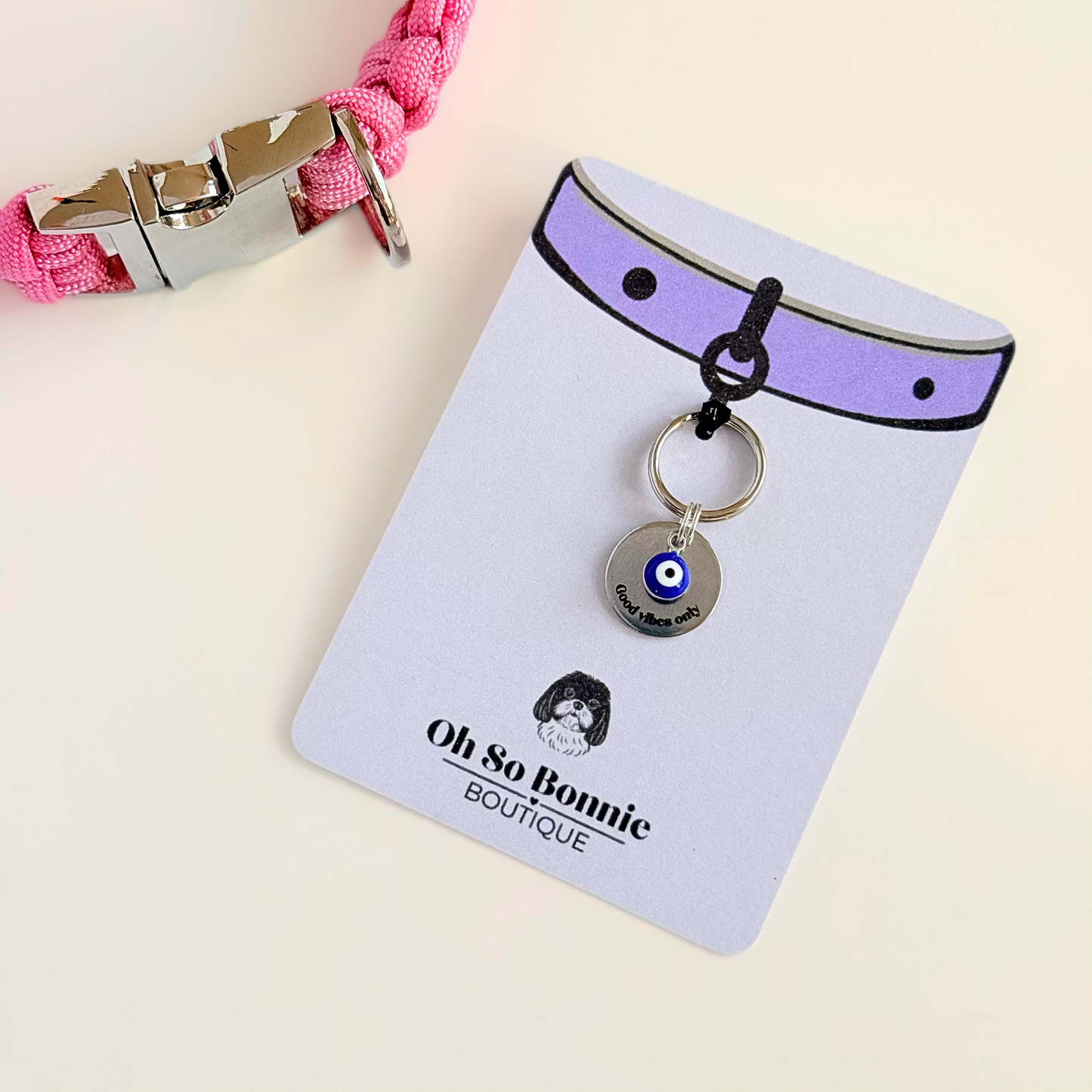 Silver plated brass and stainless steel 15mm dog collar charm with ID tag. Personalised custom identification tag with a small, blue Turkish Evil Eye charm, on a 15mm silver split ring. Engraved with the words ‘Good vibes only’ on the front. Pictured on the display card next to a pink paracord dog collar.