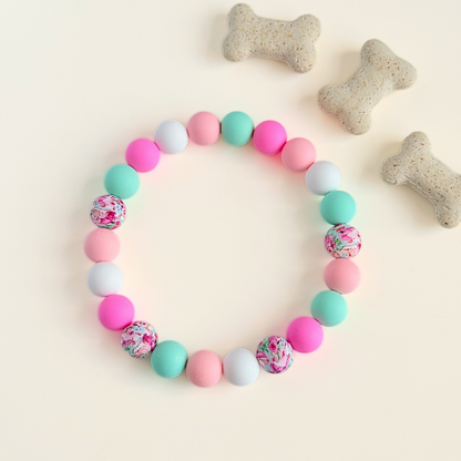 Silicone beaded dog necklace made with 15mm round beads in pink, green, white and a floral flowers print. On a strong, elastic stretch cord. Pictured from above, next to some dog biscuits.￼