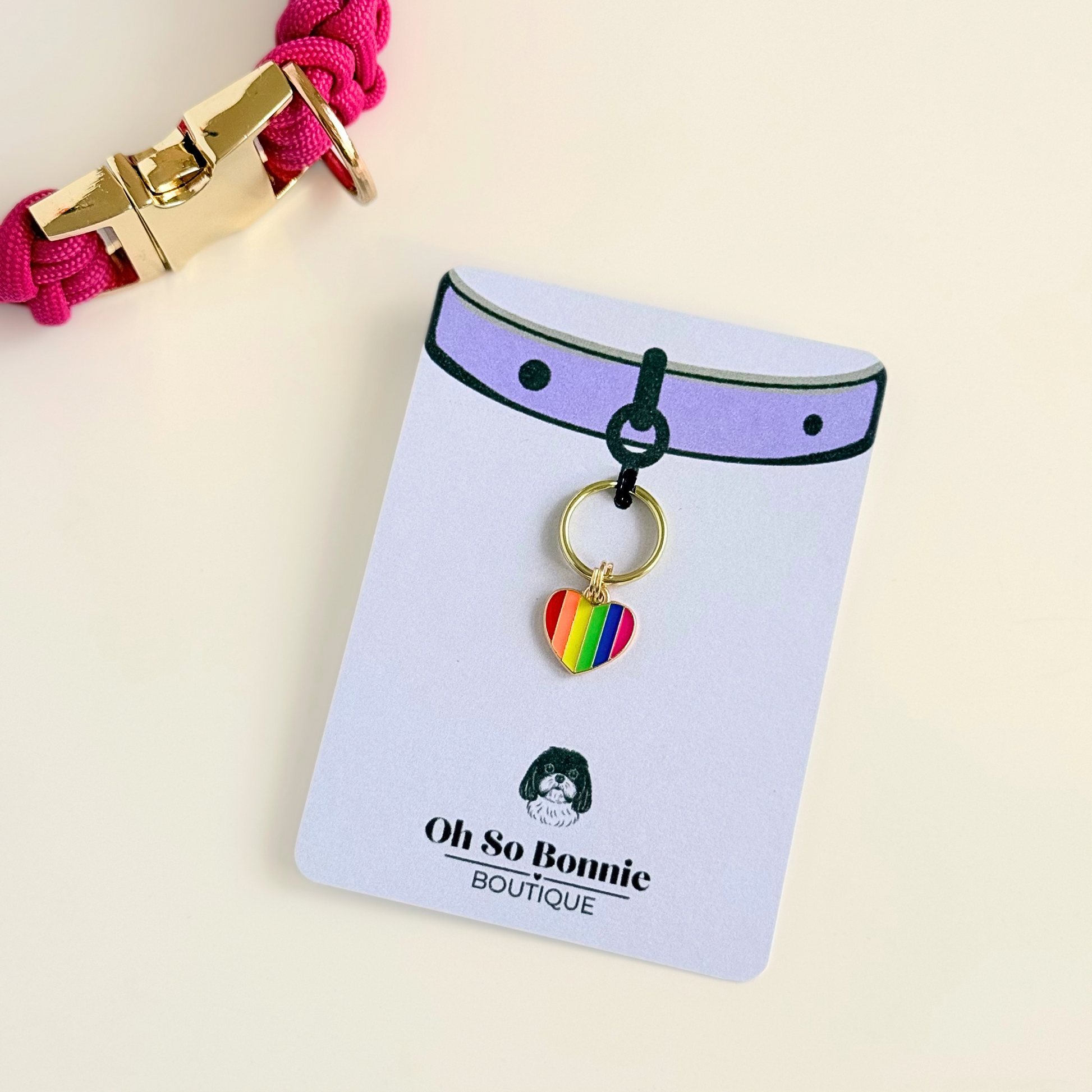 Rainbow striped heart shaped dog collar charm made with an enamel charm and a gold plated brass split ring. Pictured on the display card, next to a pink paracord dog collar.