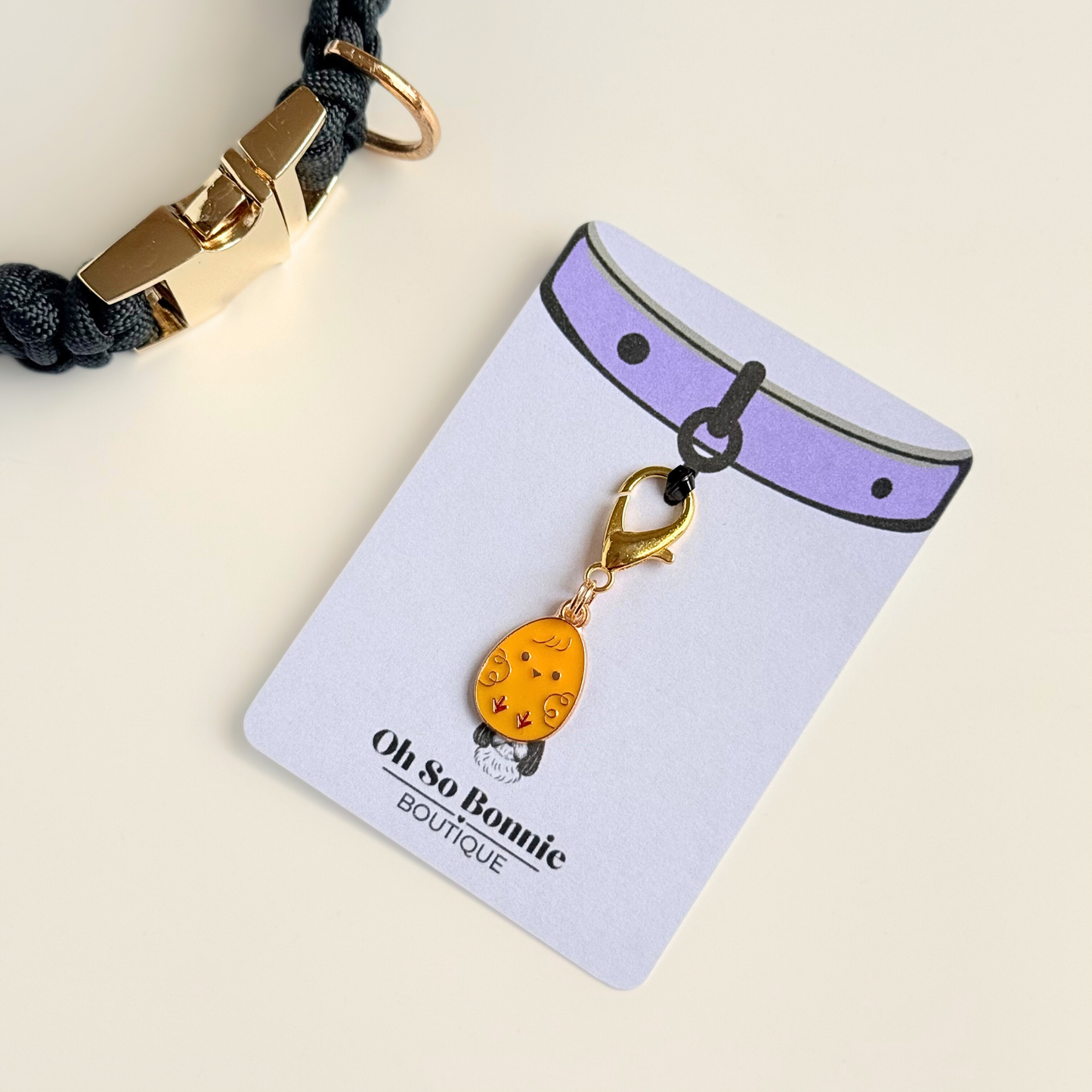 Yellow chick Easter egg enamel dog collar charm that easily attaches to collars and harnesses with a gold plated lobster clasp. Pictured on the display card next to a black paracord dog collar.￼