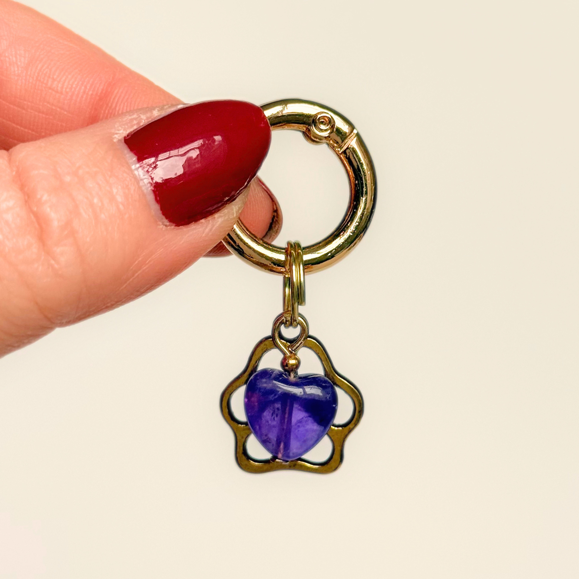 Crystal for dogs pet collar charm featuring a 10mm purple amethyst heart shaped bead and a brass gold coloured paw print tag. Pictured being held against a cream background.