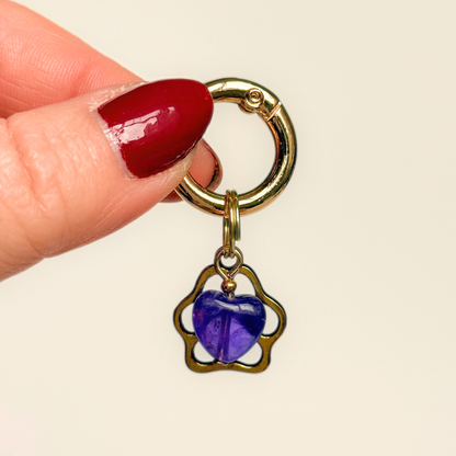 Crystal for dogs pet collar charm featuring a 10mm purple amethyst heart shaped bead and a brass gold coloured paw print tag. Pictured being held against a cream background.
