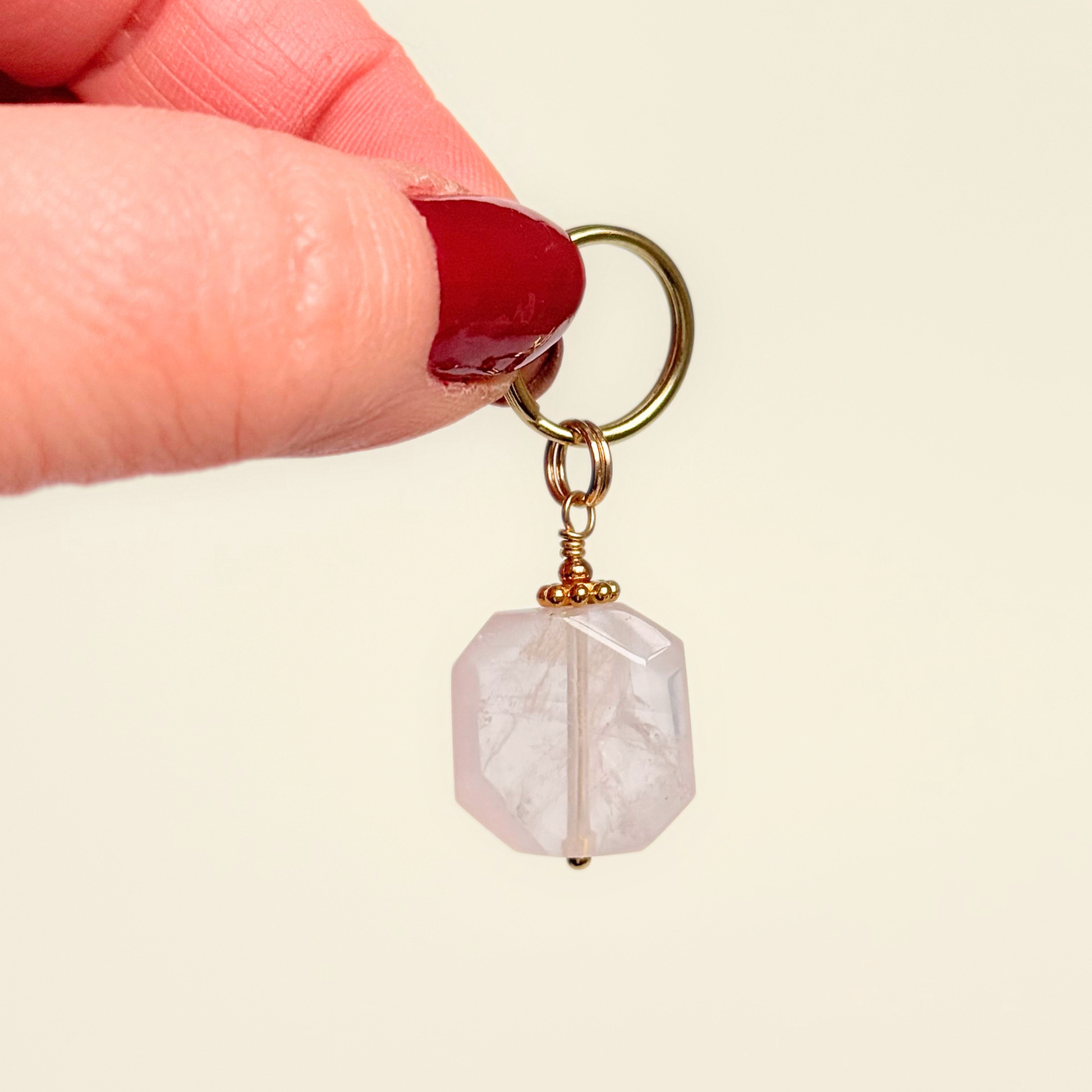 Handmade dog collar charm featuring a 17mm pink rose quartz natural gemstone faceted square crystal bead. Hung on a gold plated brass split ring with a daisy gold spacer bead that attaches onto your pet collar. Rose quartz can help pets with love, comfort and calming. Pictured being held against a cream background.