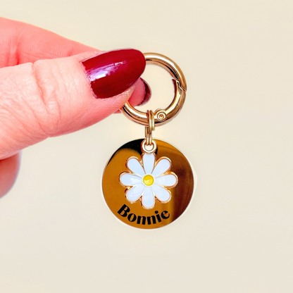 Gold white daisy flower dog collar ID tag, Enamel floral charm with dog name and address details. easily clips onto collar or harness. Pictured being held against a cream background.