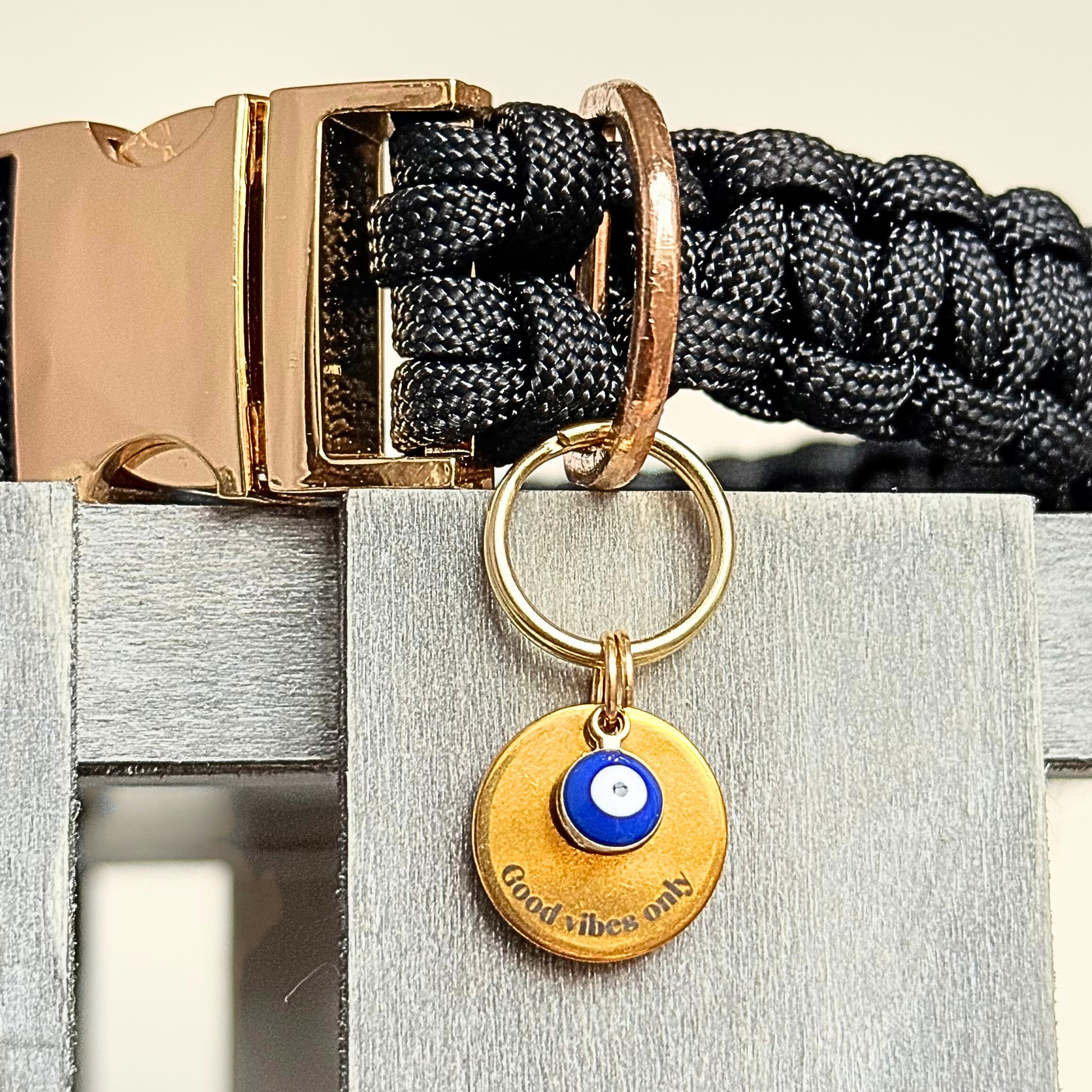 Gold plated brass and stainless steel 15mm dog collar charm with ID tag. Personalised custom identification tag with a small, blue Turkish Evil Eye charm, on a 15mm brass split ring. Engraved with the words ‘Good vibes only’ on the front. Pictured attached to a black paracord dog collar.