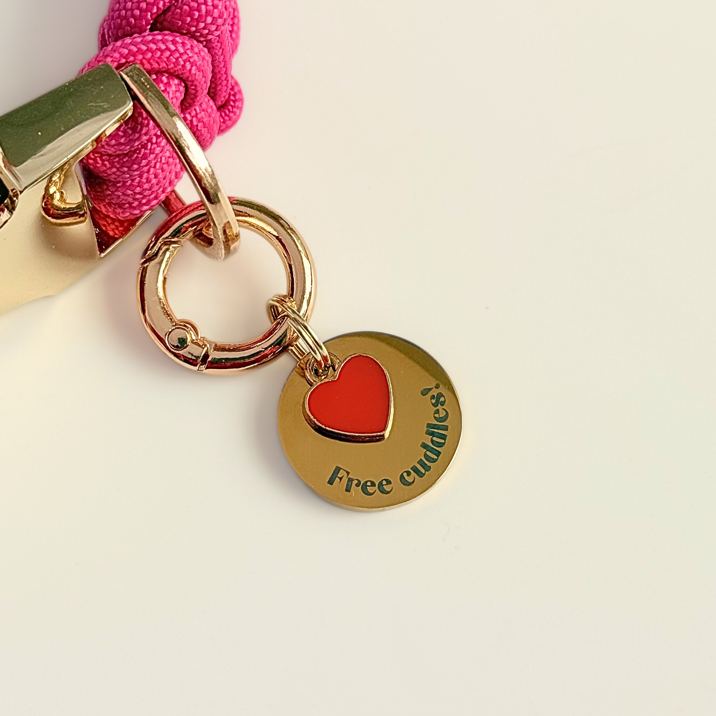 Gold plated brass and stainless steel 20mm dog collar charm with ID tag. Personalised custom identification tag with a small, red love heart charm, on a  brass carabiner style o-ring ring. Engraved with the words ‘Free cuddles’! on the front. Pictured attached to a pink paracord dog collar against a cream background.