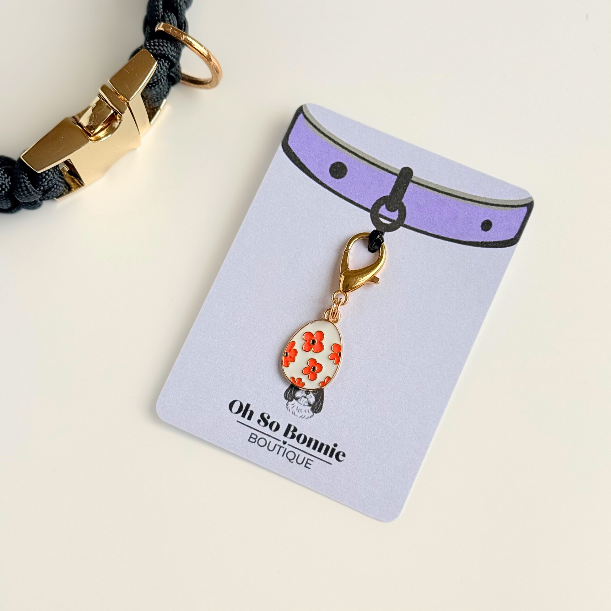 White with red flowers Easter egg enamel dog collar charm that easily attaches to collars and harnesses with a gold plated lobster clasp. Pictured on the display card next to a black paracord dog collar.￼