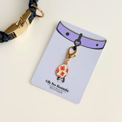 White with red flowers Easter egg enamel dog collar charm that easily attaches to collars and harnesses with a gold plated lobster clasp. Pictured on the display card next to a black paracord dog collar.￼