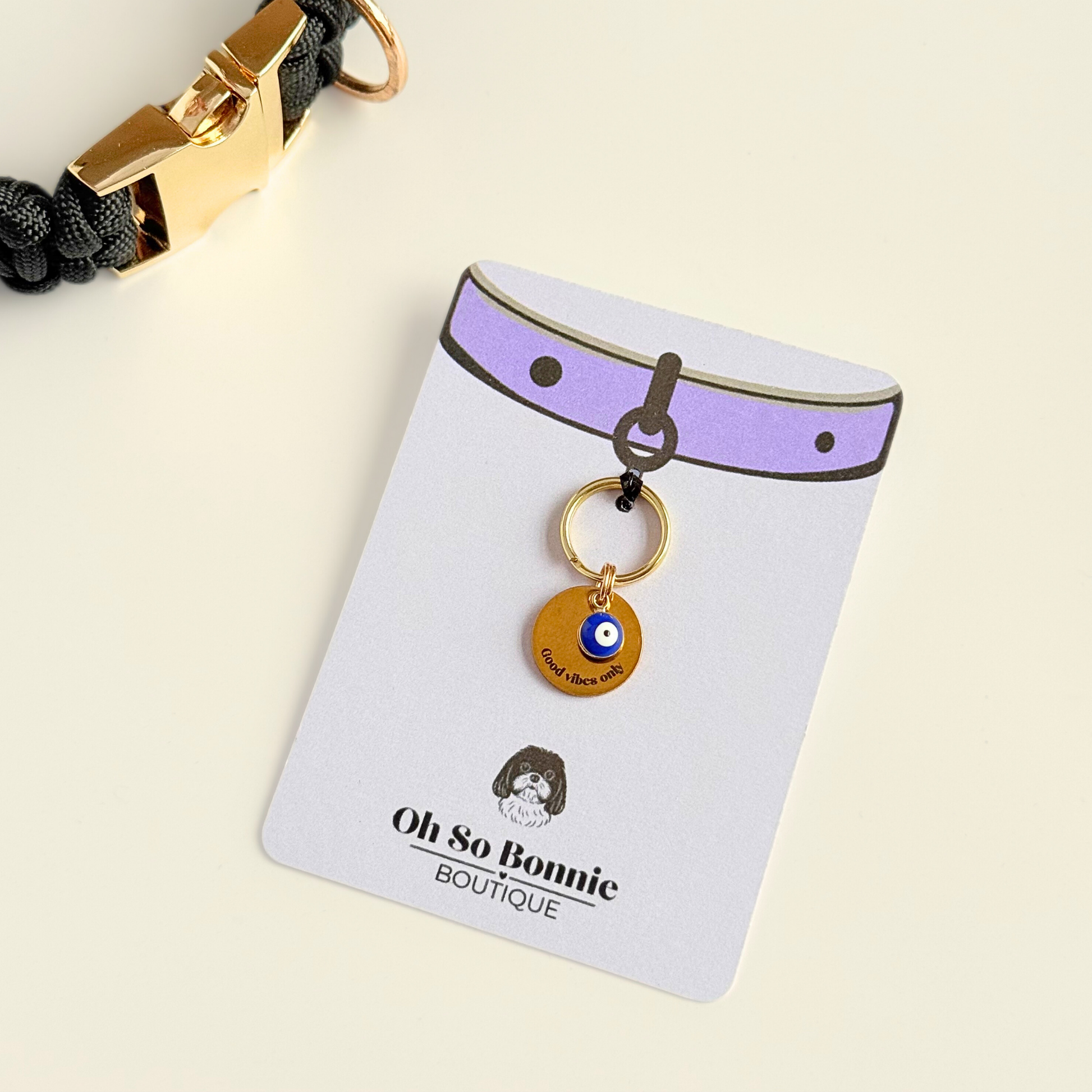 Gold plated brass and stainless steel 15mm dog collar charm with ID tag. Personalised custom identification tag with a small, blue Turkish Evil Eye charm, on a 15mm brass split ring. Engraved with the words ‘Good vibes only’ on the front. Pictured on the display card next to a black paracord dog collar.