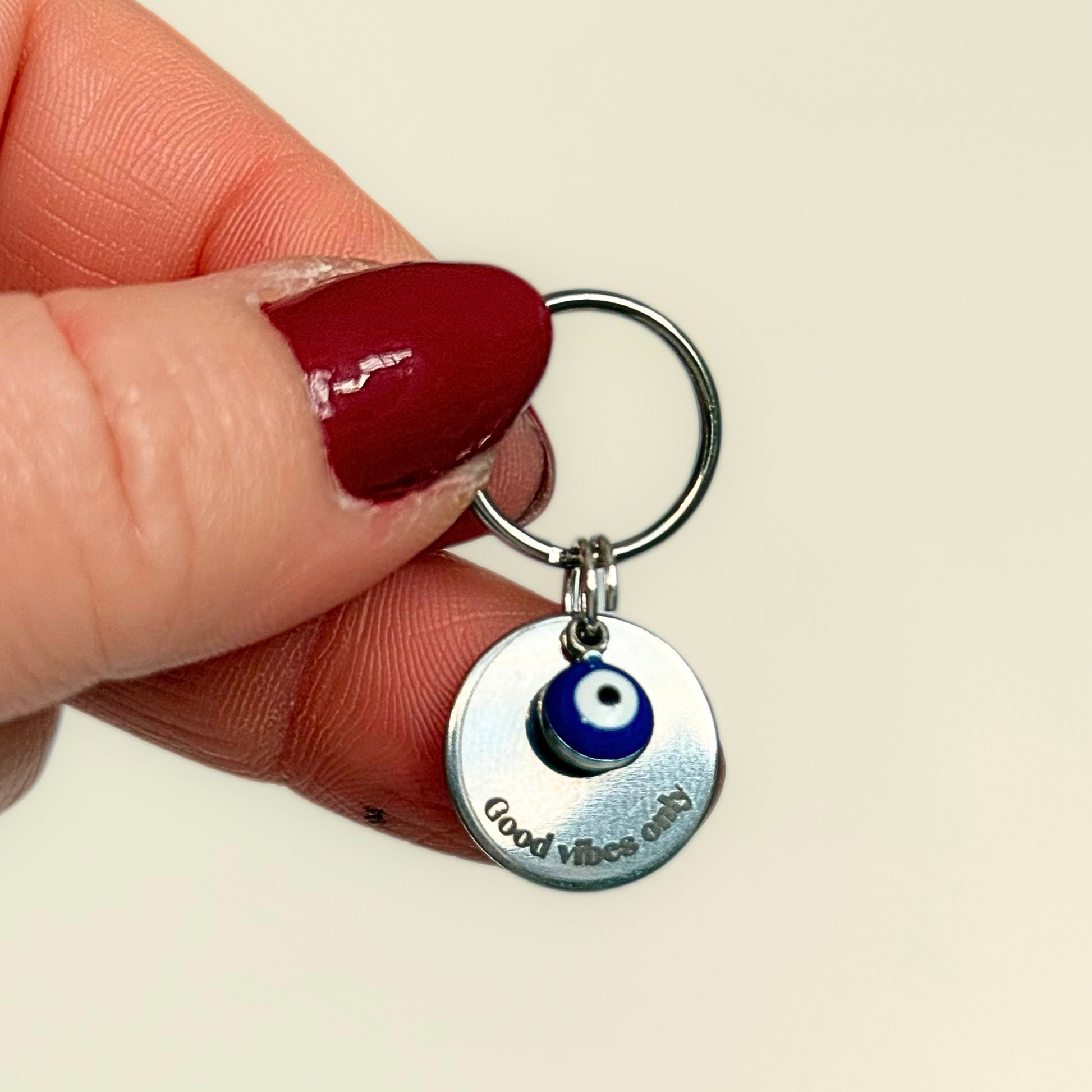 Silver plated brass and stainless steel 15mm dog collar charm with ID tag. Personalised custom identification tag with a small, blue Turkish Evil Eye charm, on a 15mm silver split ring. Engraved with the words ‘Good vibes only’ on the front. Pictured being held up against a cream background.
