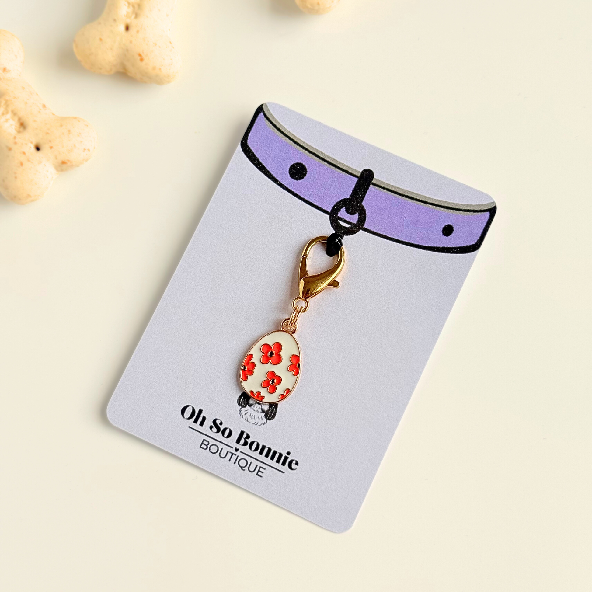 White with red flowers Easter egg enamel dog collar charm that easily attaches to collars and harnesses with a gold plated lobster clasp. Pictured on the display card next to some dog bone biscuits.￼