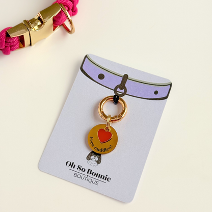 Gold plated brass and stainless steel 20mm dog collar charm with ID tag. Personalised custom identification tag with a small, red love heart charm, on a  brass carabiner style o-ring. Engraved with the words ‘Free cuddles’! on the front. Pictured on the display card next to a pink paracord dog collar.