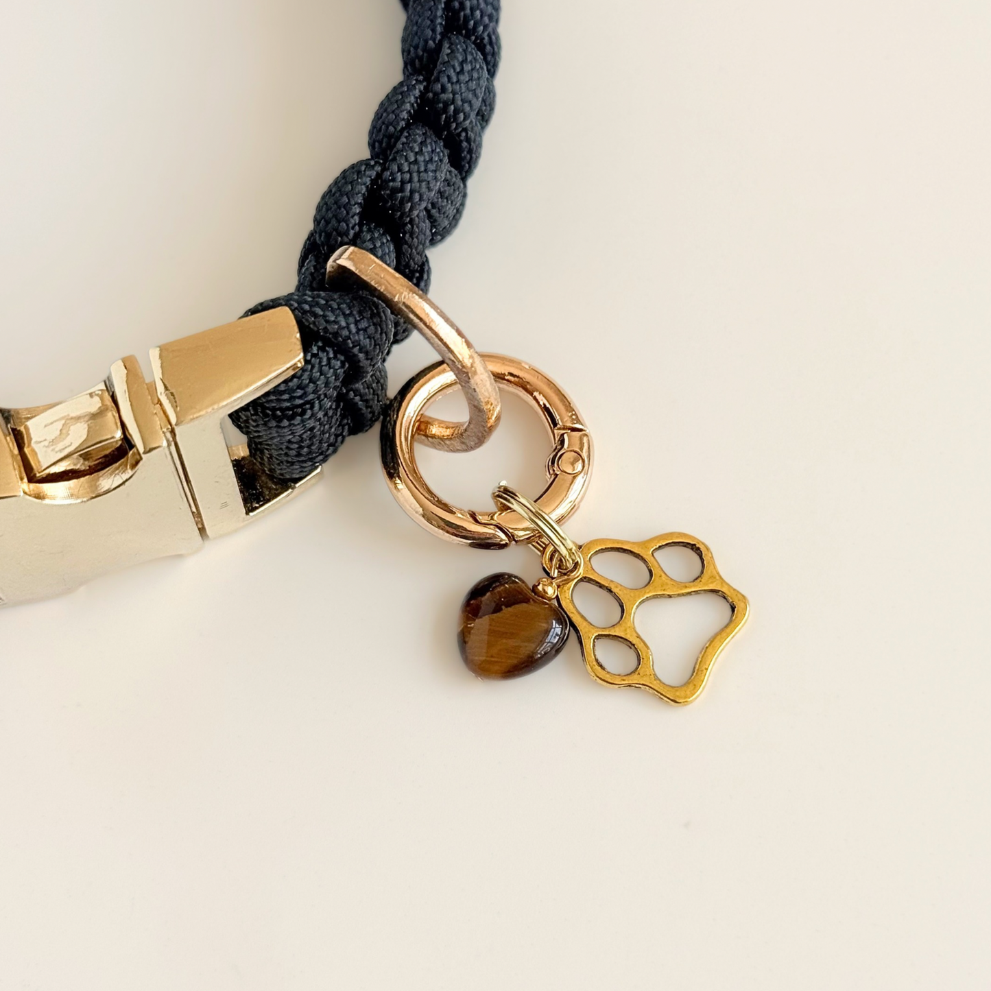 Crystal for dogs pet collar charm featuring a 10mm tigers eye heart shaped bead and a brass gold coloured paw print tag. Pictured attached to a black paracord dog collar.
