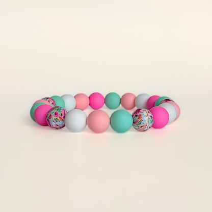 Silicone beaded dog necklace made with 15mm round beads in pink, green, white and a floral flowers print. On a strong, elastic stretch cord. Pictured against a cream background.￼