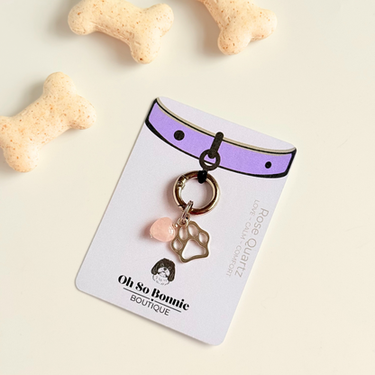 Crystal for dogs pet collar charm featuring a 10mm rose quartz heart shaped bead and a brass silver coloured paw print tag. A stone for love, calm and comfort. Pictured on the display card, next to some dog bone biscuits