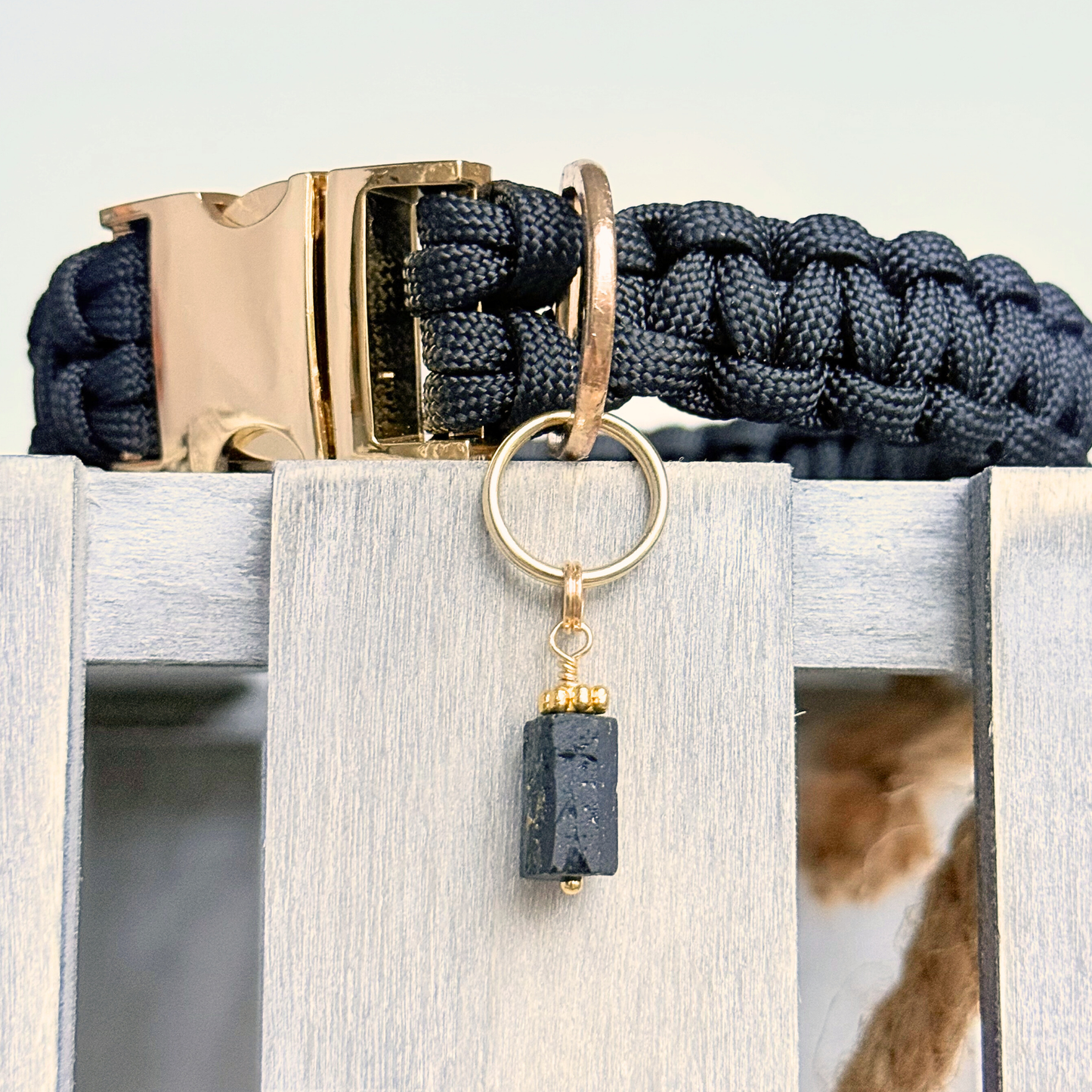 Crystal dog collar charm featuring a raw black tourmaline natural bead with gold plated accents. Featuring a brass split ring for easy attaching to your pet’s collar. Close up picture of the charm attached to a black paracord dog collar.