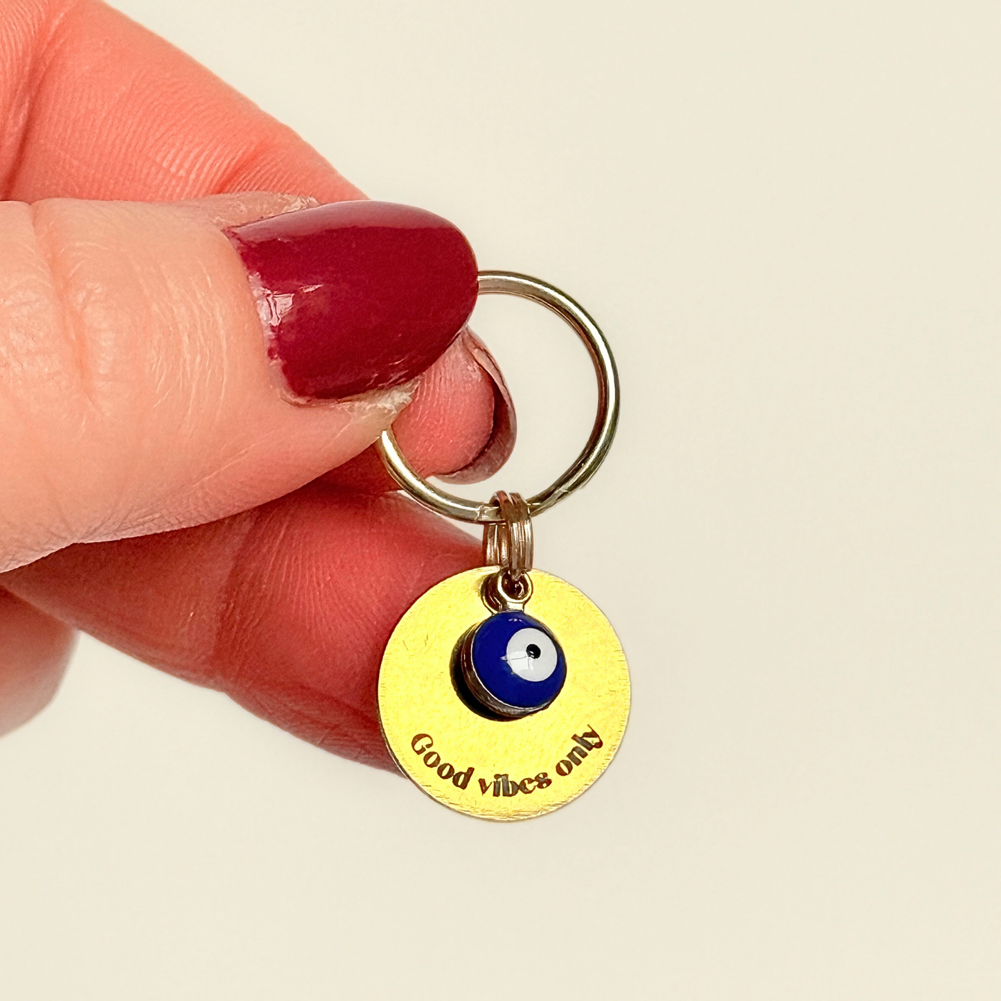 Gold plated brass and stainless steel 15mm dog collar charm with ID tag. Personalised custom identification tag with a small, blue Turkish Evil Eye charm, on a 15mm brass split ring. Engraved with the words ‘Good vibes only’ on the front. Pictured being held up against a cream background.