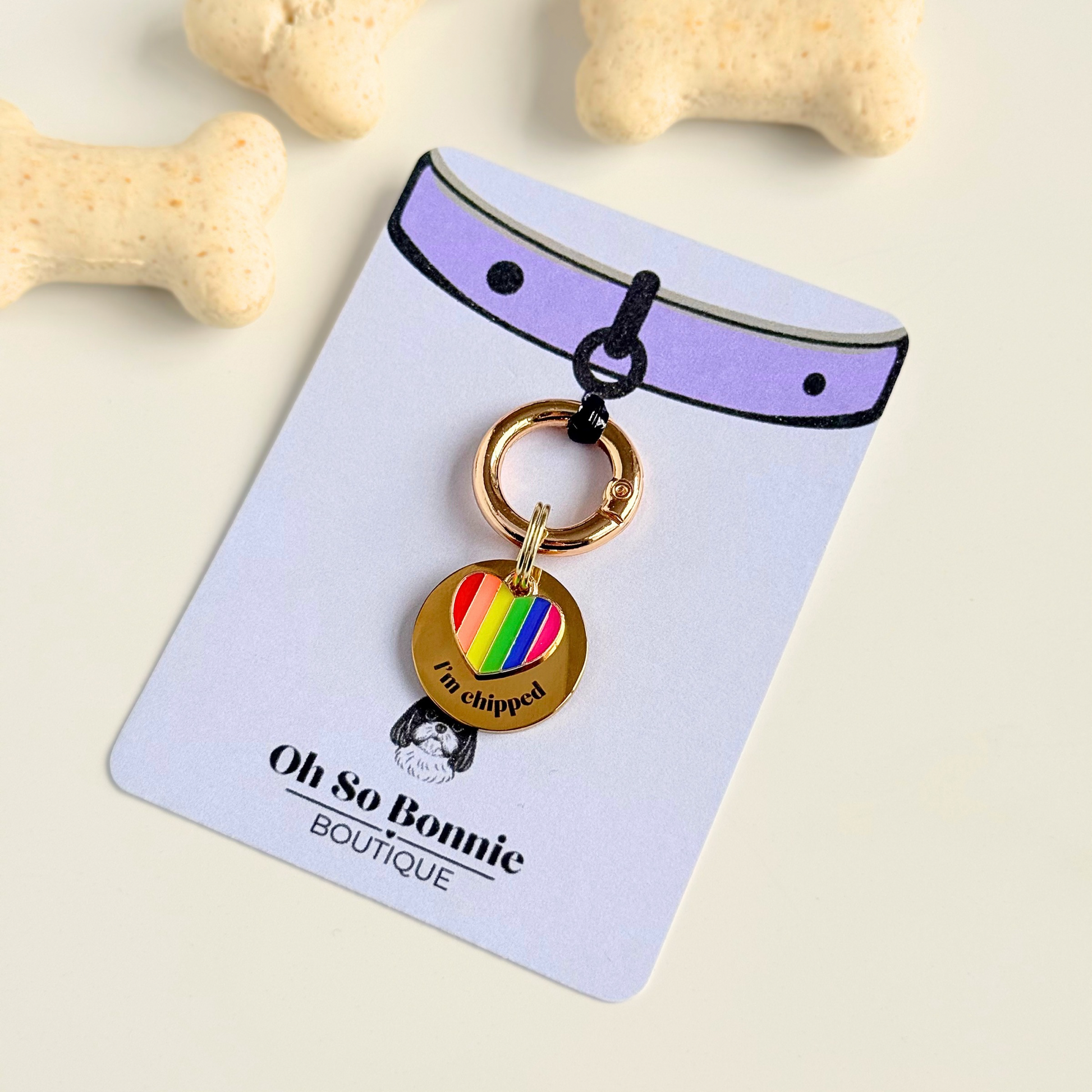 Gold brass rainbow striped heart dog collar ID tag, Enamel charm with dog name and address details. easily clips onto collar or harness. Pictured on the display card next to some dog biscuits.