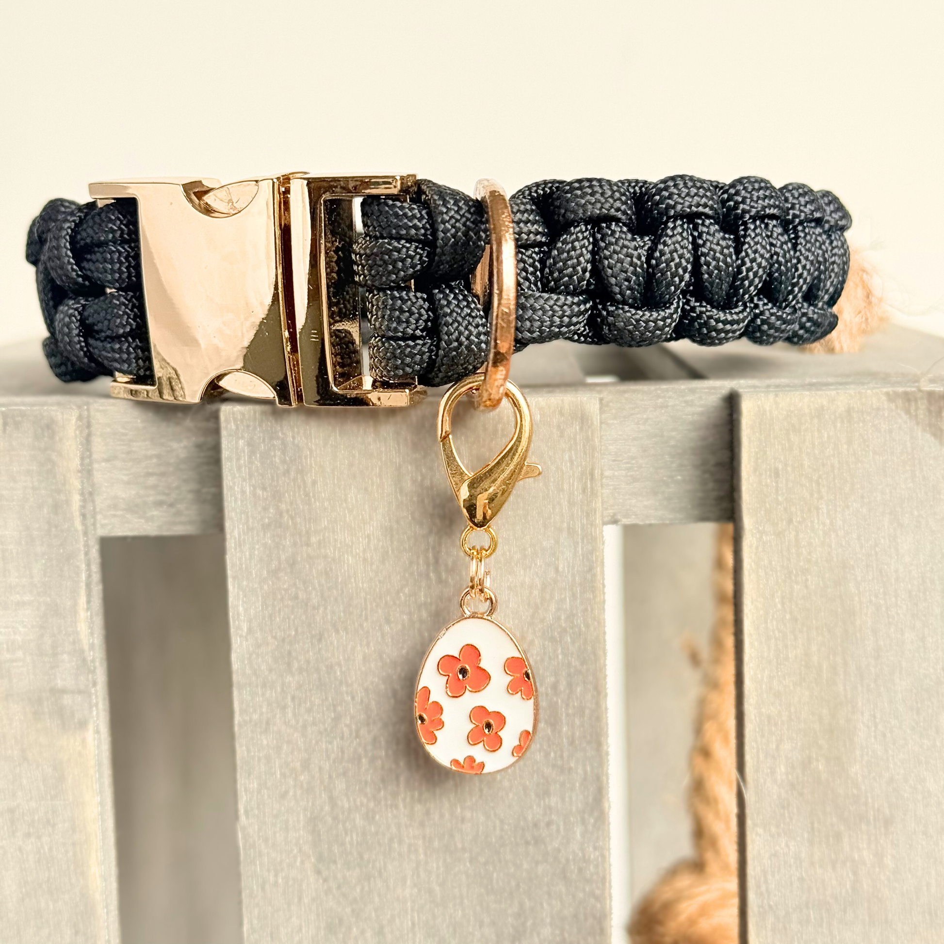 White with red flowers Easter egg enamel dog collar charm that easily attaches to collars and harnesses with a gold plated lobster clasp. Close-up picture attached to a black paracord dog collar.￼