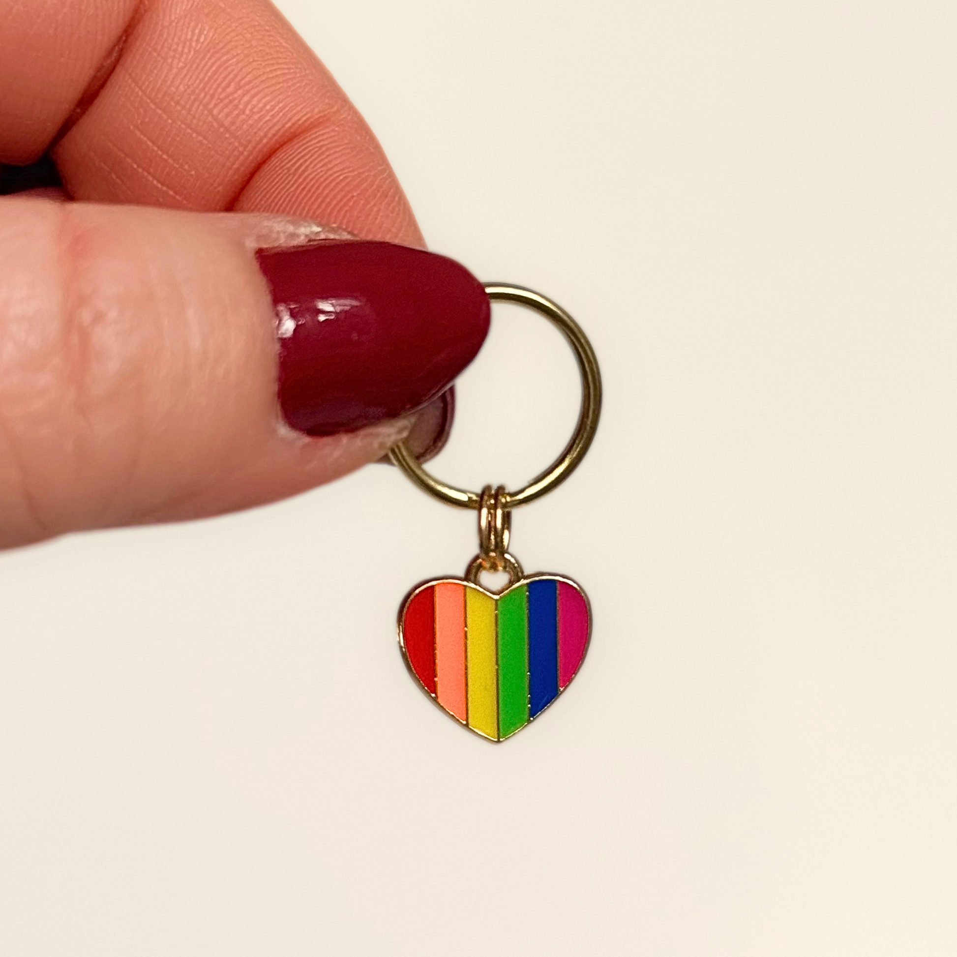 Rainbow striped heart shaped dog collar charm made with an enamel charm and a gold plated brass split ring. Pictured being held against a cream background.