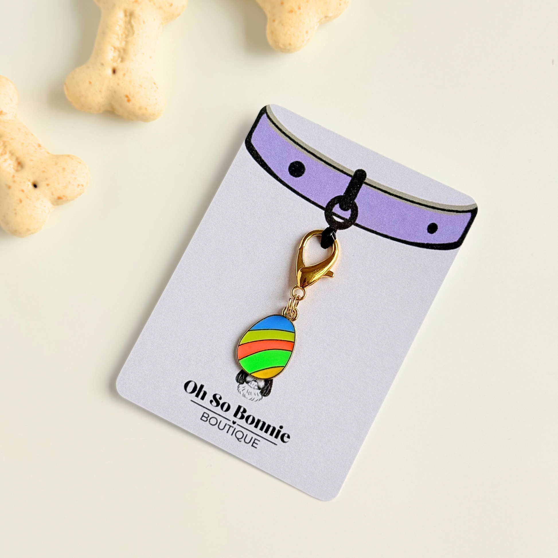 Colourful striped Easter egg enamel dog collar charm that easily attaches to collars and harnesses with a gold plated lobster clasp. Pictured on the display card next to some dog biscuits.￼