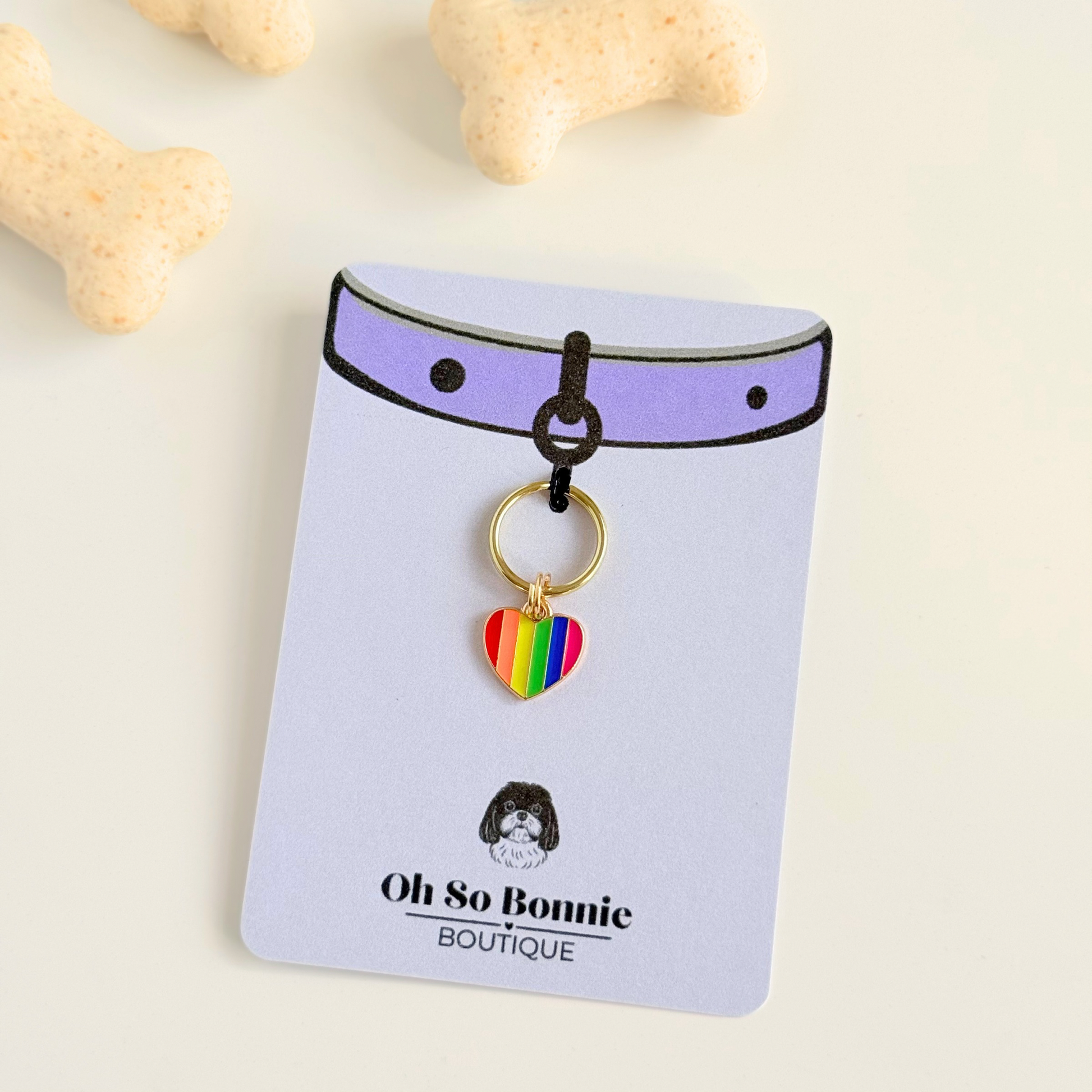Rainbow striped heart shaped dog collar charm made with an enamel charm and a gold plated brass split ring. Pictured on the display card, next to some dog biscuits.