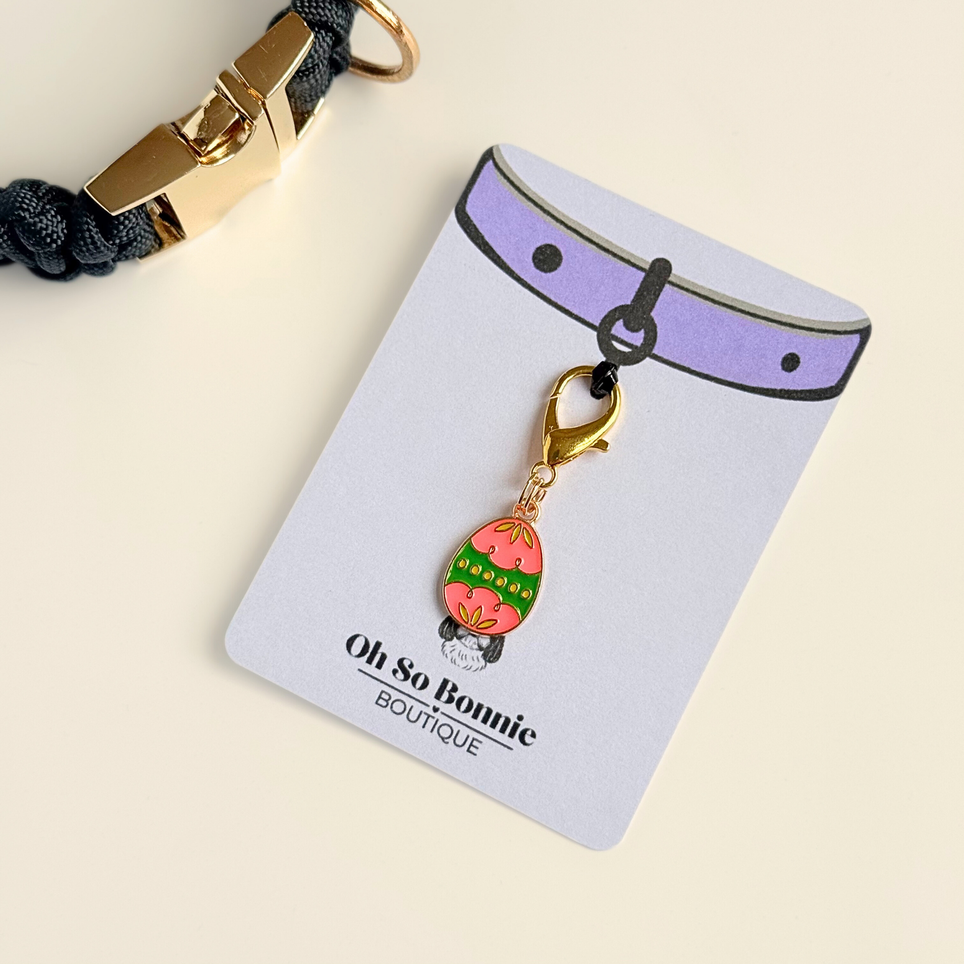 Colourful pink and green patterned Easter egg enamel dog collar charm that easily attaches to collars and harnesses with a gold plated lobster clasp.  Pictured on the display card next to a black paracord dog collar.￼