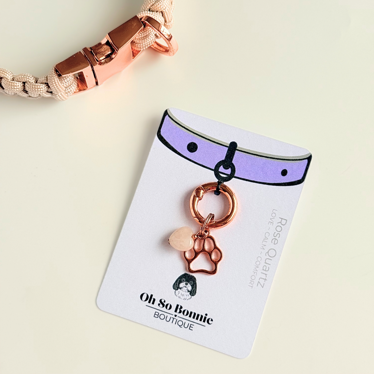 Crystal for dogs pet collar charm featuring a 10mm rose quartz heart shaped bead and a brass rose-gold coloured paw print tag. A stone for love, calm and comfort. Pictured on the display card next to a pearl rose collar.