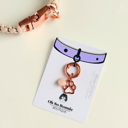 Crystal for dogs pet collar charm featuring a 10mm rose quartz heart shaped bead and a brass rose-gold coloured paw print tag. A stone for love, calm and comfort. Pictured on the display card next to a pearl rose collar.