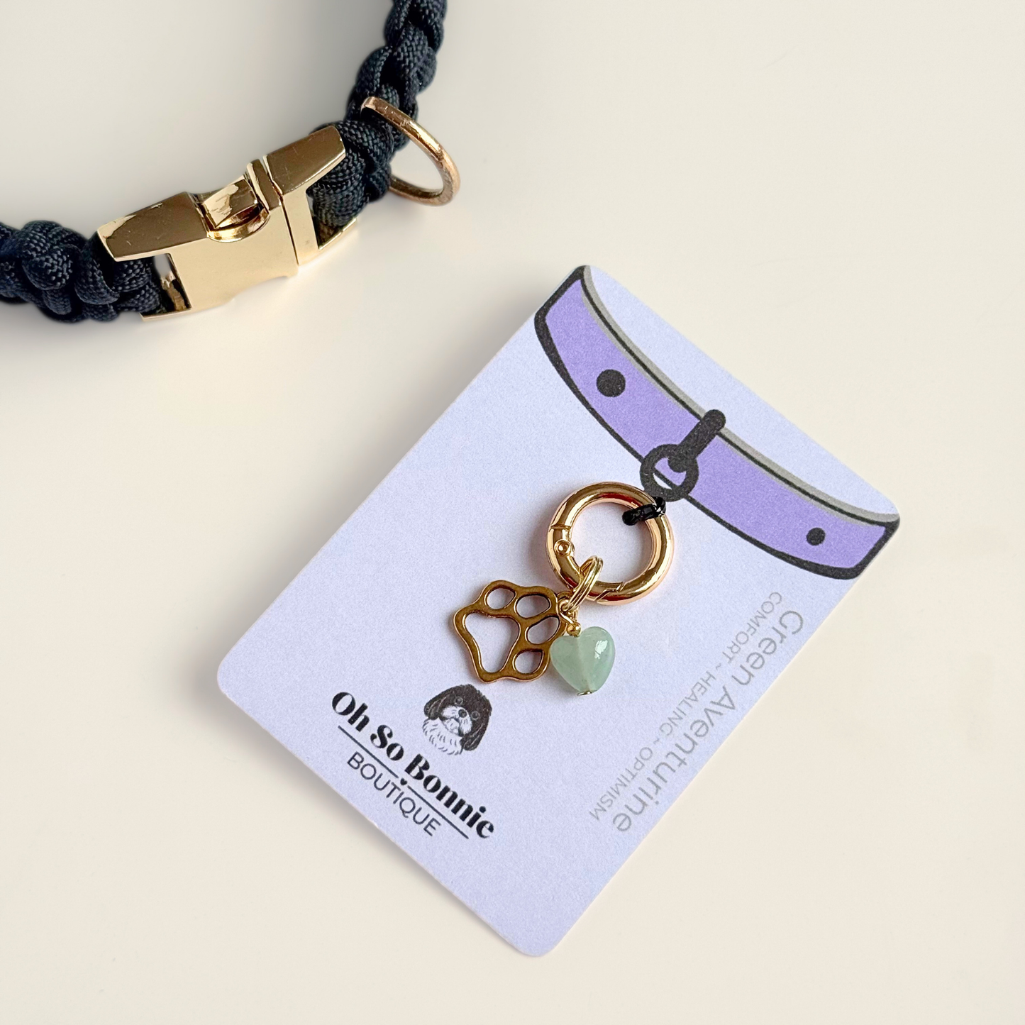 Crystal for dogs pet collar charm featuring a 10mm green aventurine heart shaped bead and a brass gold coloured paw print tag. A stone for comfort, healing and optimism. Pictured on the display card next to a black paracord dog collar.
