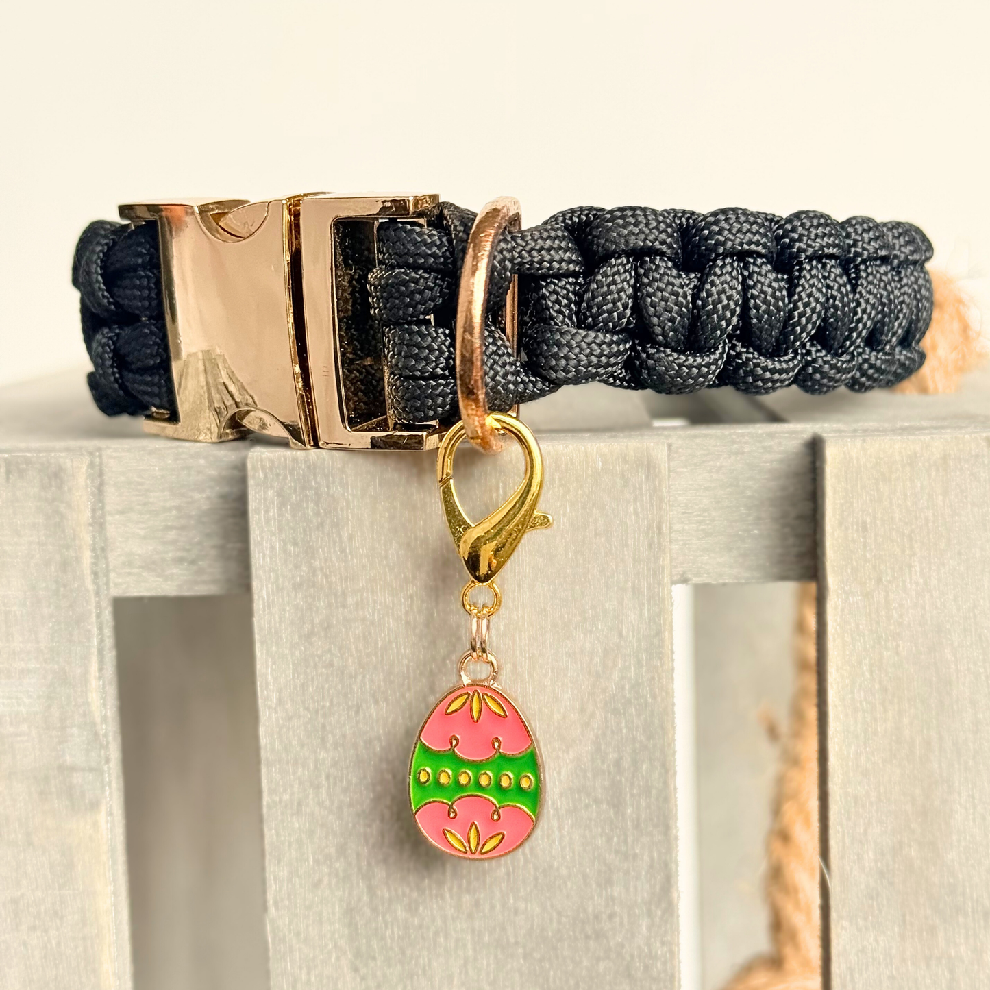 Colourful pink and green patterned Easter egg enamel dog collar charm that easily attaches to collars and harnesses with a gold plated lobster clasp. Close-up picture of it attached to a black paracord dog collar.￼
