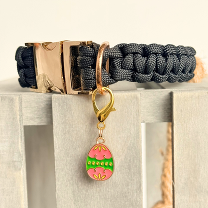 Colourful pink and green patterned Easter egg enamel dog collar charm that easily attaches to collars and harnesses with a gold plated lobster clasp. Close-up picture of it attached to a black paracord dog collar.￼