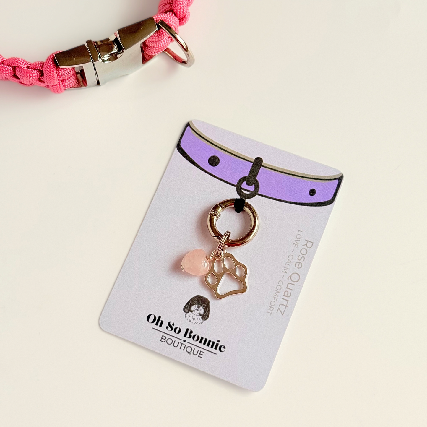 Crystal for dogs pet collar charm featuring a 10mm rose quartz heart shaped bead and a brass silver coloured paw print tag. A stone for love, calm and comfort. Pictured on the display card next to a pink collar.