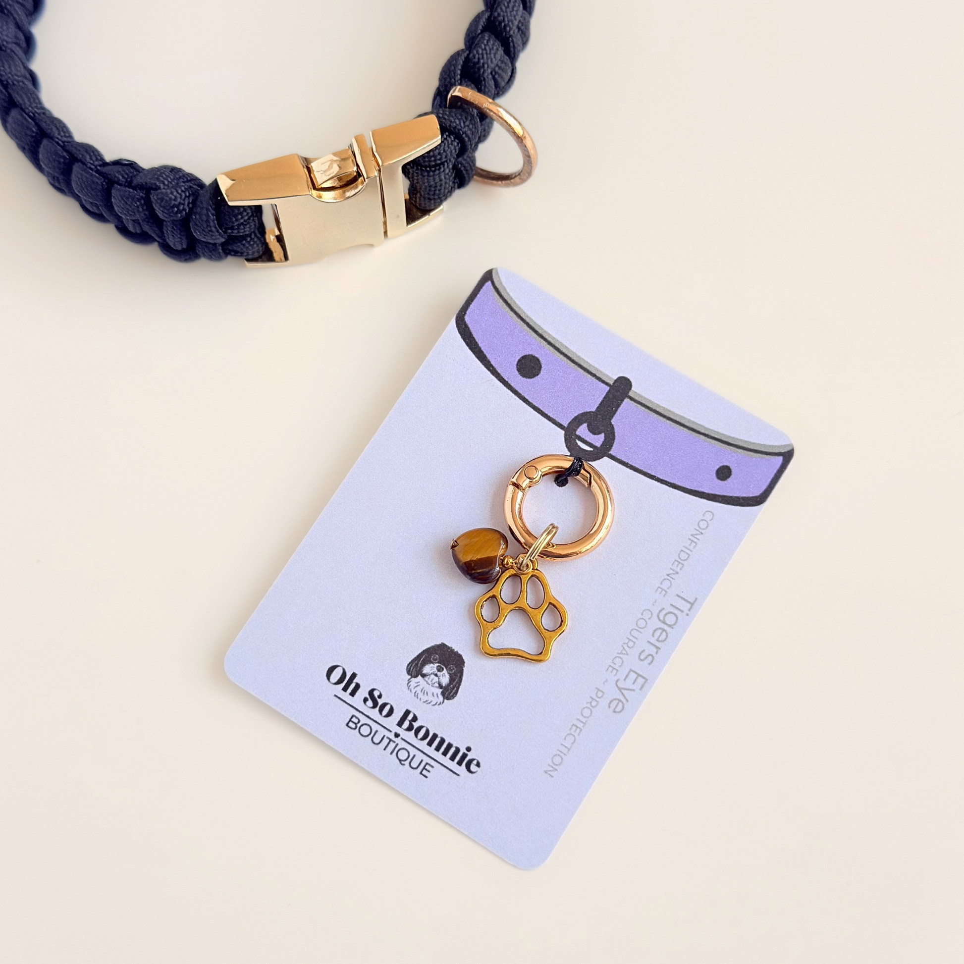 Crystal for dogs pet collar charm featuring a 10mm tigers eye heart shaped bead and a brass gold coloured paw print tag. Pictured on the display card, next to a black paracord dog collar.