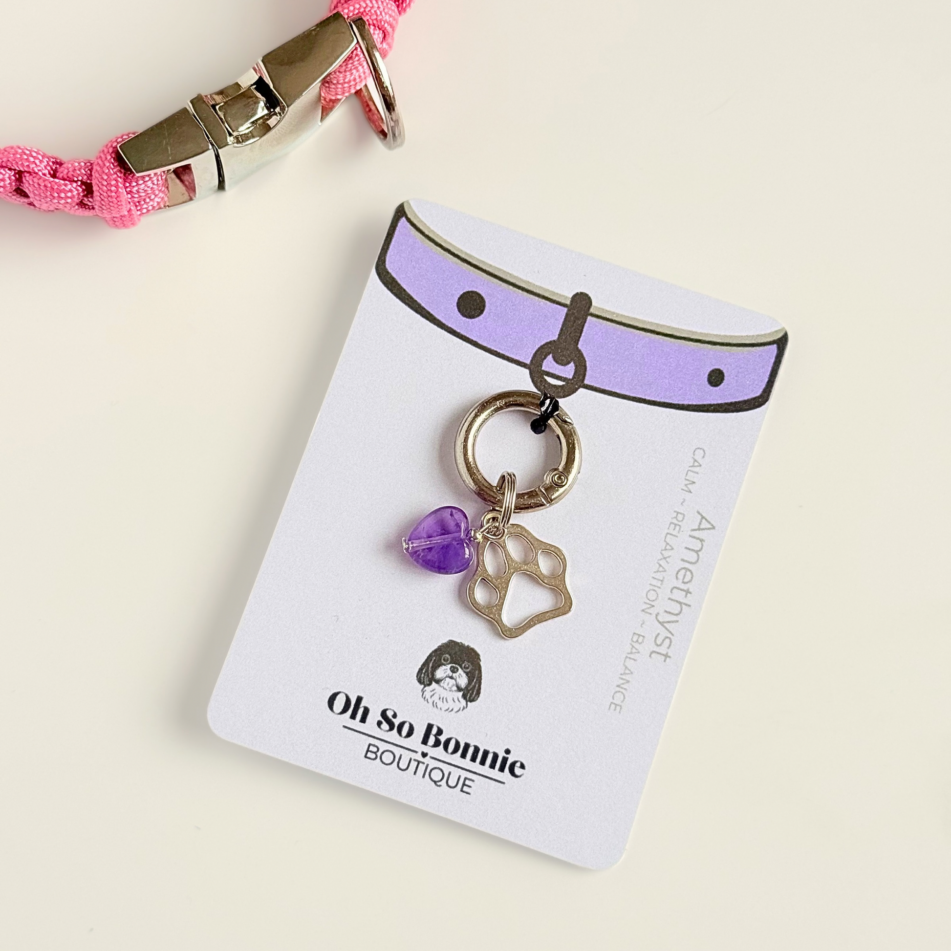 Crystal for dogs pet collar charm featuring a 10mm purple amethyst heart shaped bead and a brass silver coloured paw print tag. Pictured on the isplay card, next to a pink paracord dog collar.