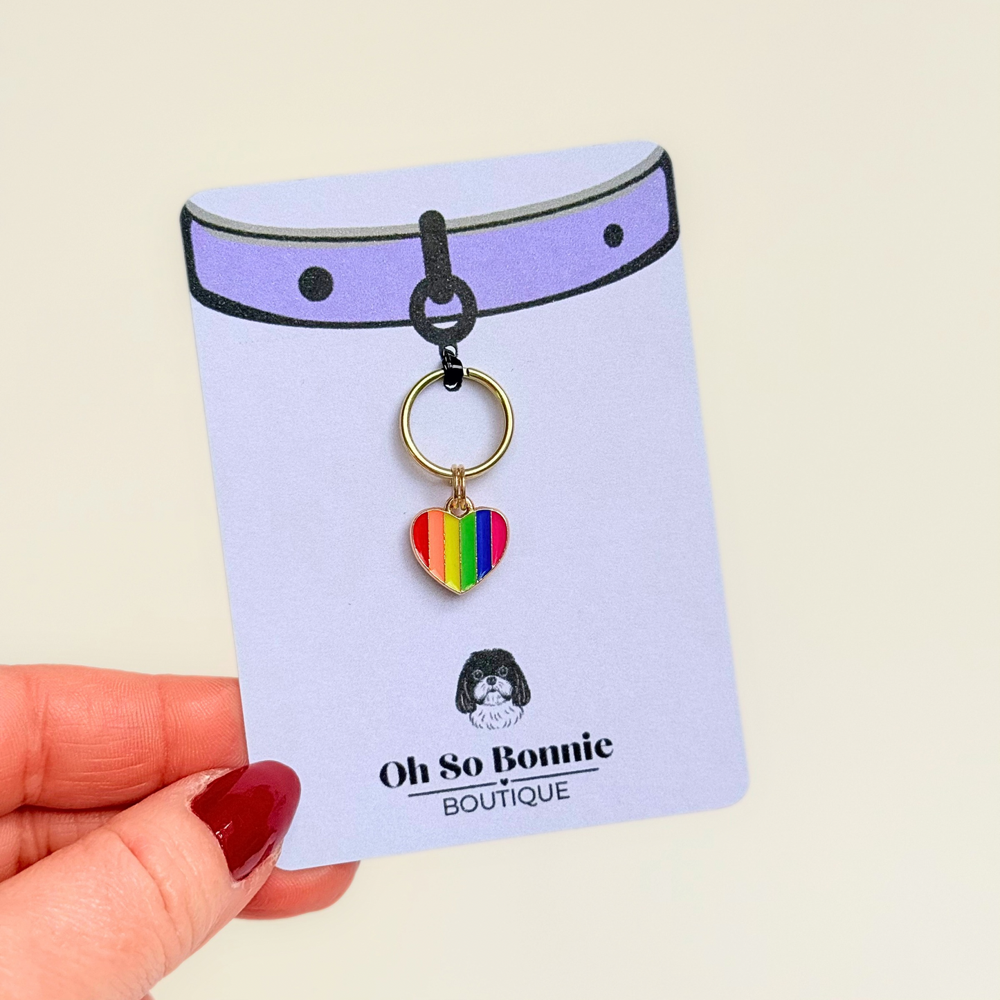 Rainbow striped heart shaped dog collar charm made with an enamel charm and a gold plated brass split ring. Pictured being held on the display card.