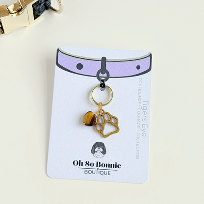 Crystal for dogs pet collar charm featuring a 10mm tigers eye heart shaped bead and a brass gold coloured paw print tag. Pictured on the display card, next to a black paracord dog collar.