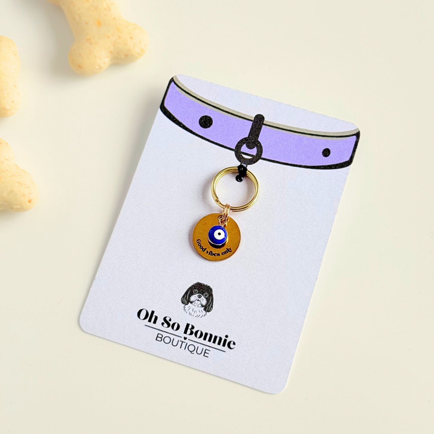 Gold plated brass and stainless steel 15mm dog collar charm with ID tag. Personalised custom identification tag with a small, blue Turkish Evil Eye charm, on a 15mm brass split ring. Engraved with the words ‘Good vibes only’ on the front. Pictured against a cream background on the display card.
